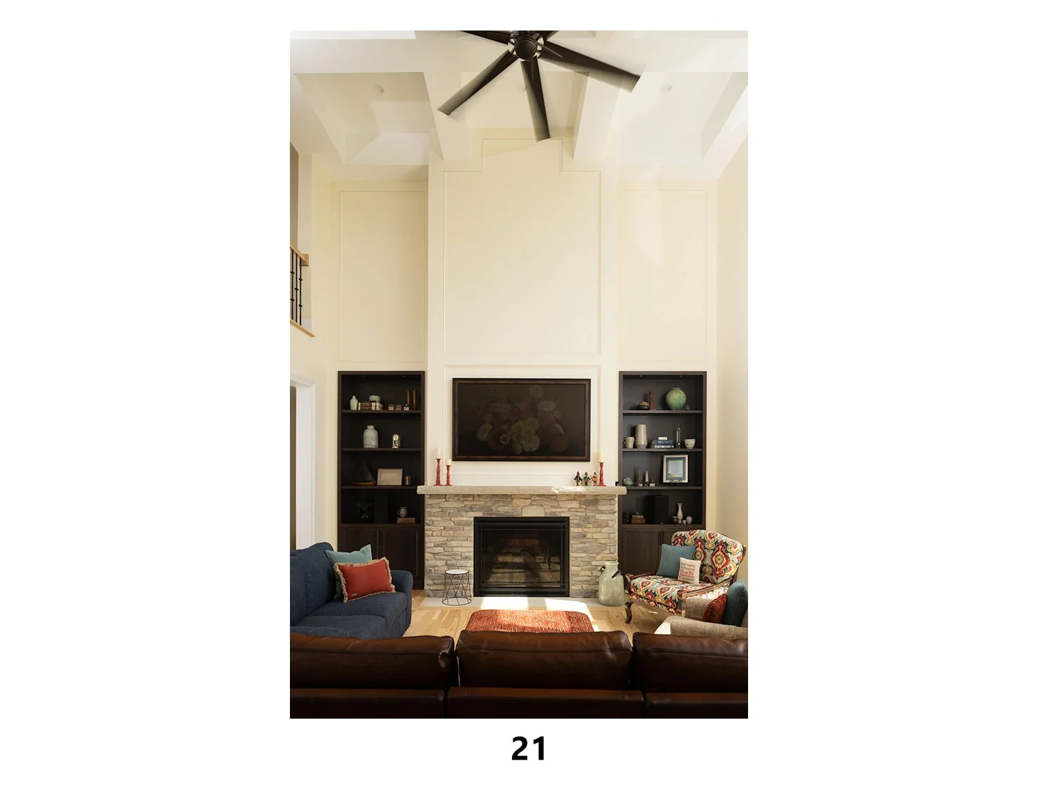Living room with a stone fireplace, built-in shelves, a large television, ceiling fan, and seating including a blue sofa and floral armchair.