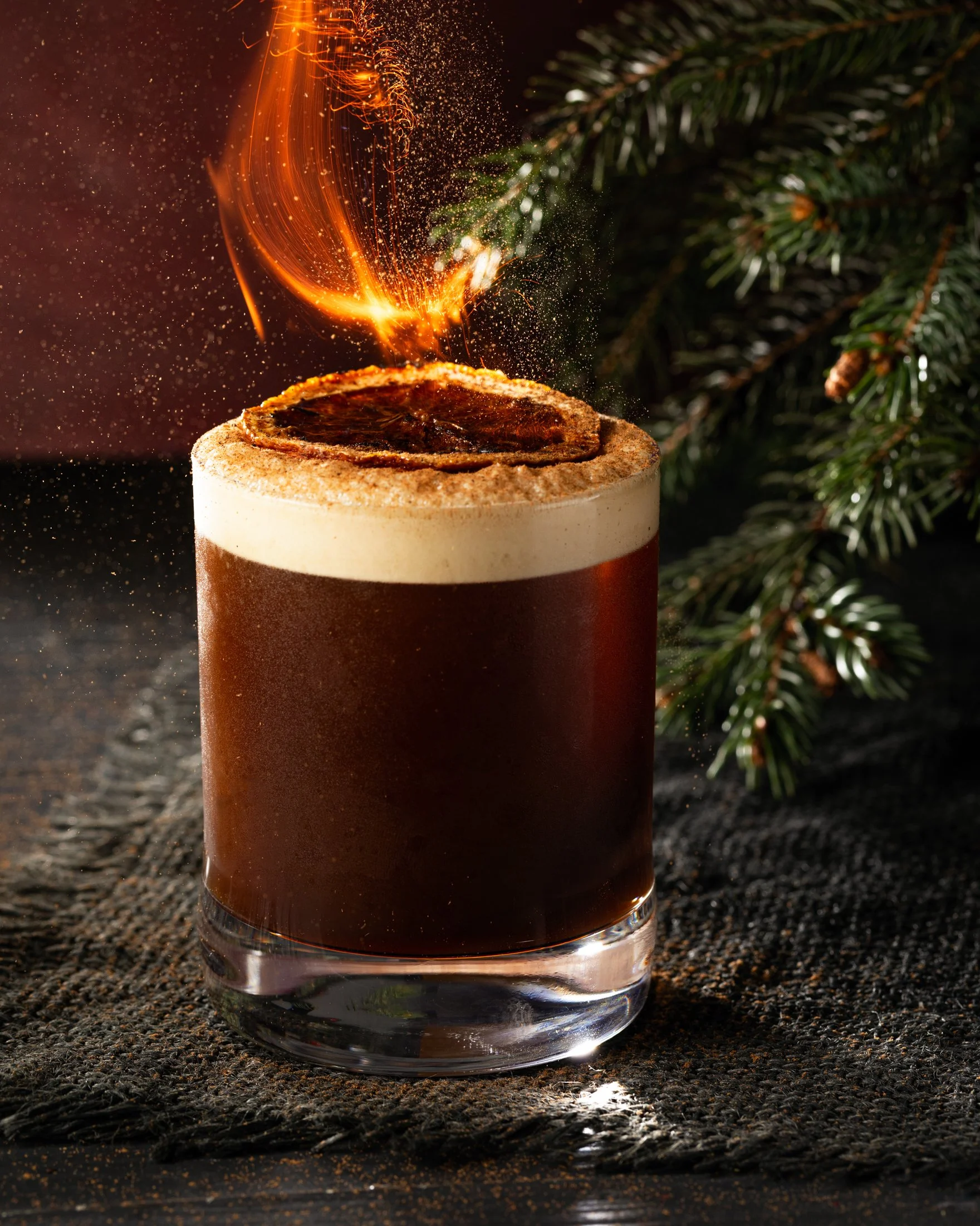 A glass of dark stout beer with a frothy head, topped with a burning cinnamon stick, set on a dark textured surface with pine branches in the background.