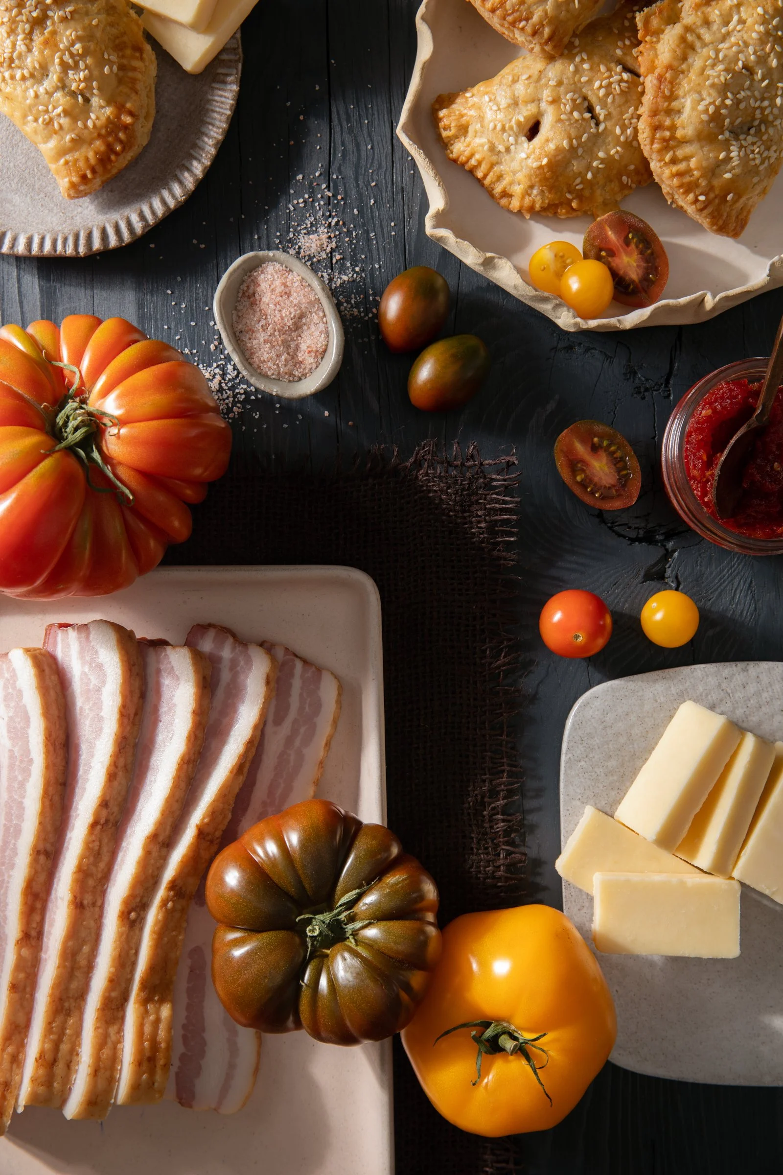 A flat lay of ingredients for a meal, including heirloom tomatoes, bacon, cheese, a jar of tomato sauce, and pastries on a dark table.