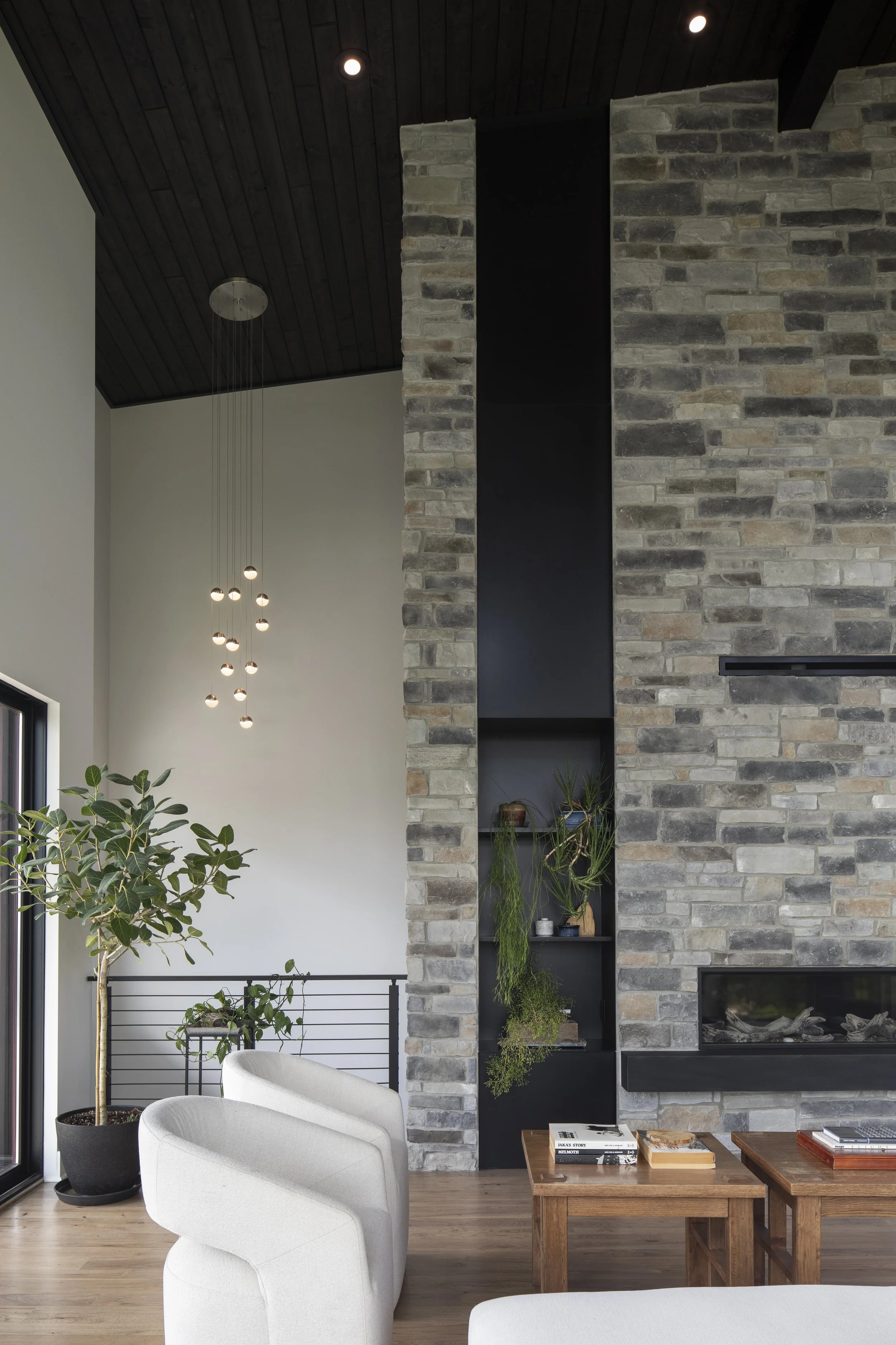 Modern living room with a tall brick fireplace, black shelving with plants, white armchairs, and a wooden coffee table, next to a large potted plant, in a room with dark wood ceiling panels.