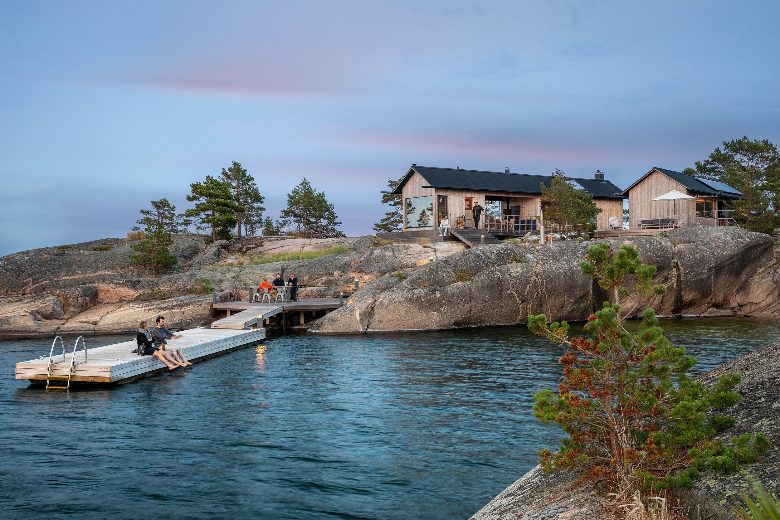 A lakeside house with a deck on rocky terrain, surrounded by trees, with a dock extending into the water, and people relaxing and socializing outdoors at dusk.