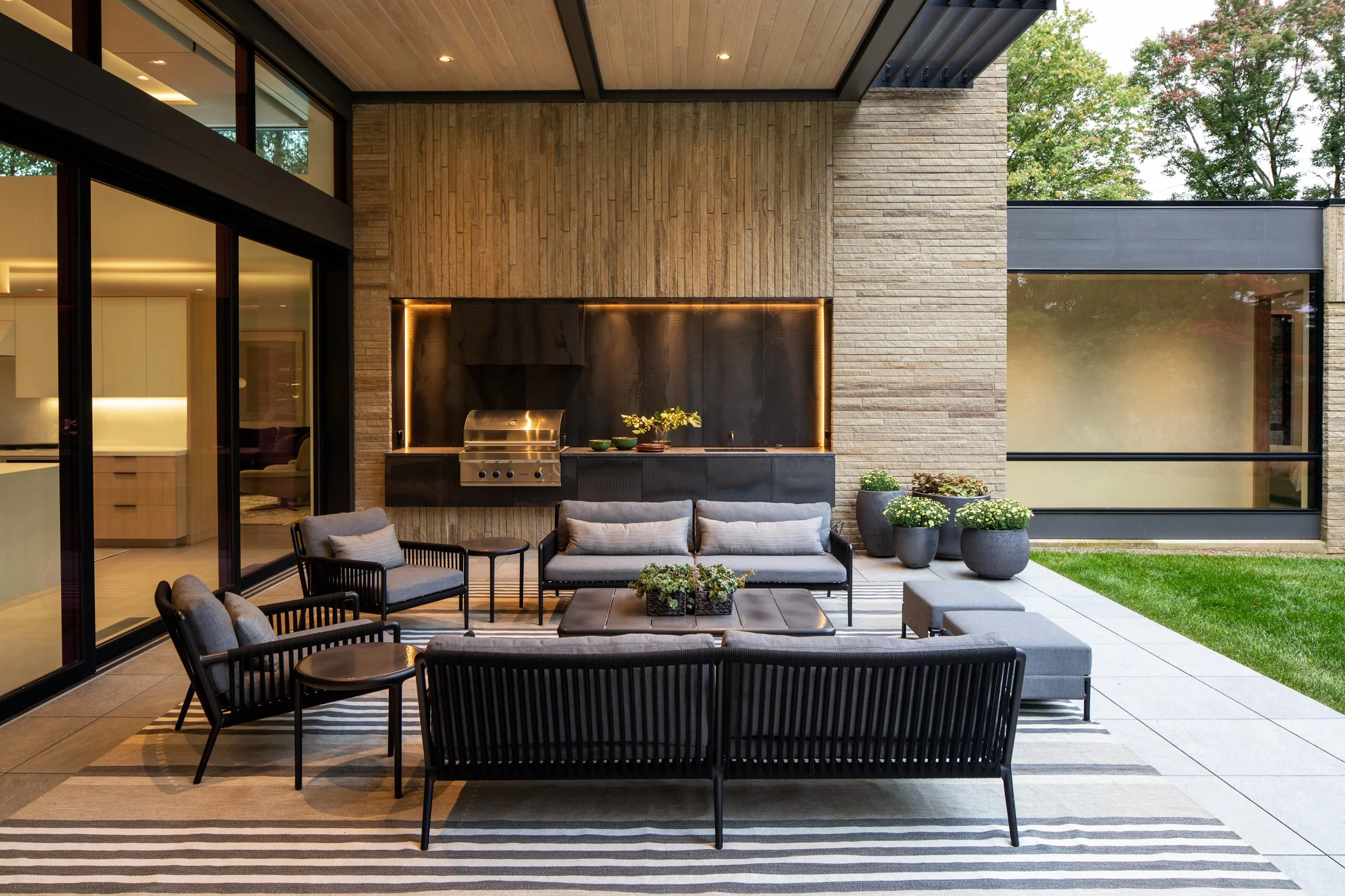 Modern outdoor patio with seating, potted plants, and built-in grill area, adjacent to a glass-walled living space, with trees in the background.