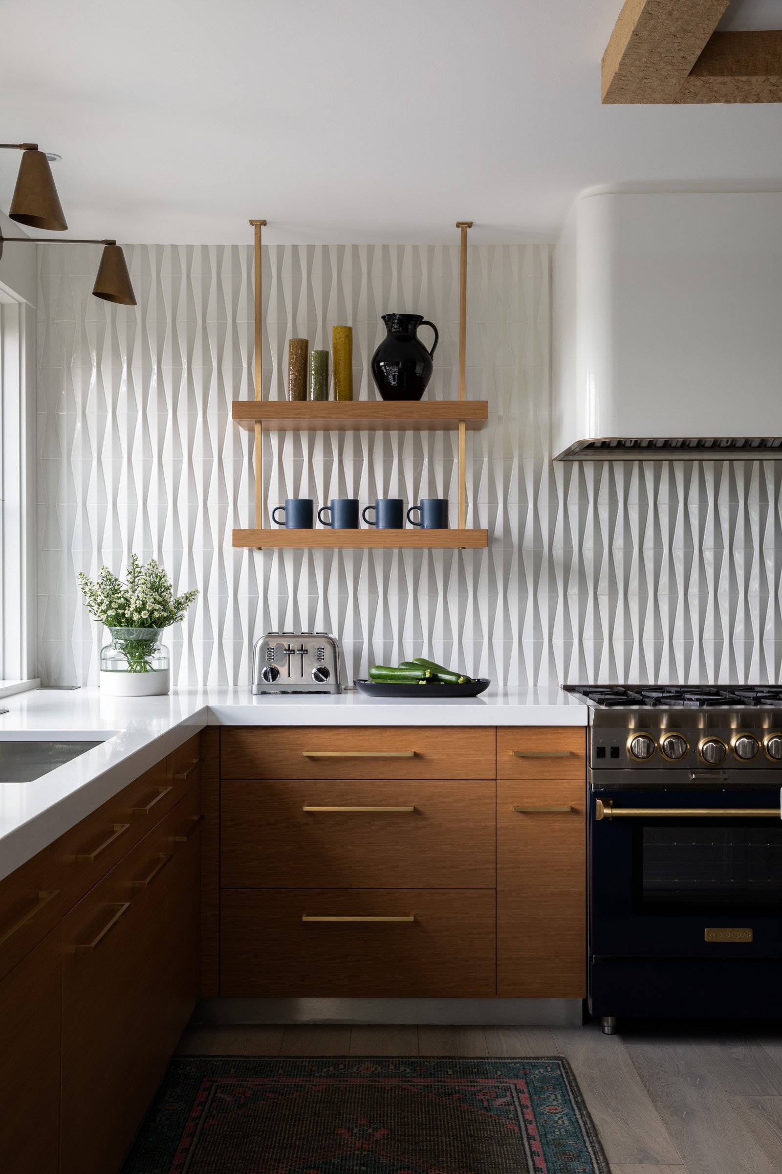 Modern kitchen with wooden cabinets, white countertop, patterned accent wall, open wooden shelves with vases and mugs, gas stove, and a potted plant. Interior design photography