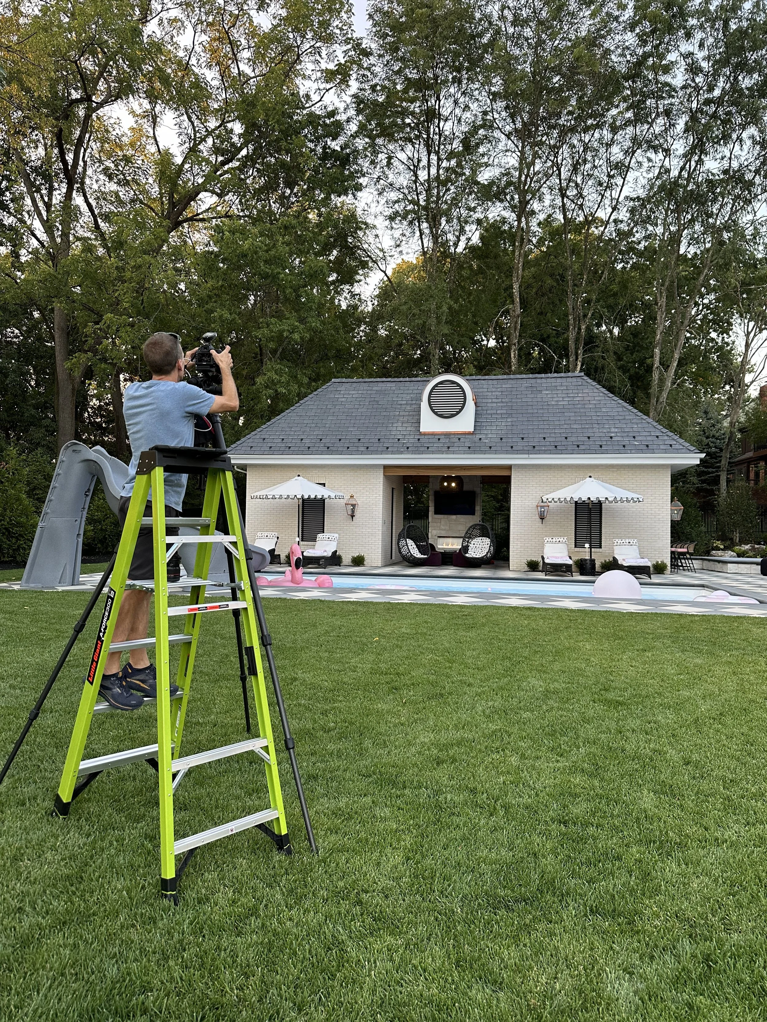 Man filming with a camera on a ladder in a backyard with a pool, poolside furniture, large umbrellas, and a small building or pool house surrounded by trees.