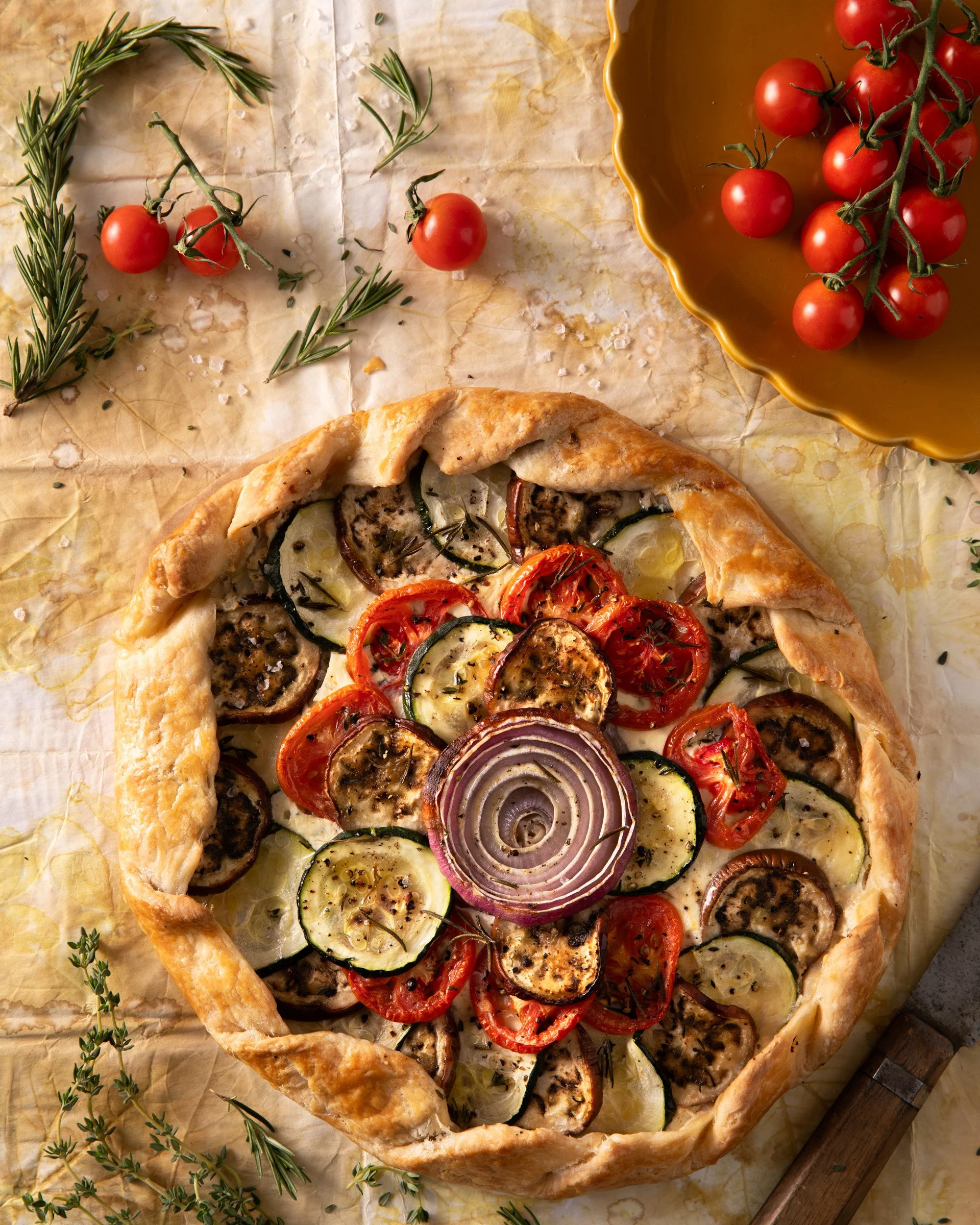 Fresh vegetable tart topped with sliced eggplant, zucchini, cherry tomatoes, and red onion, garnished with herbs on parchment paper, with cherry tomatoes and fresh herbs beside it.