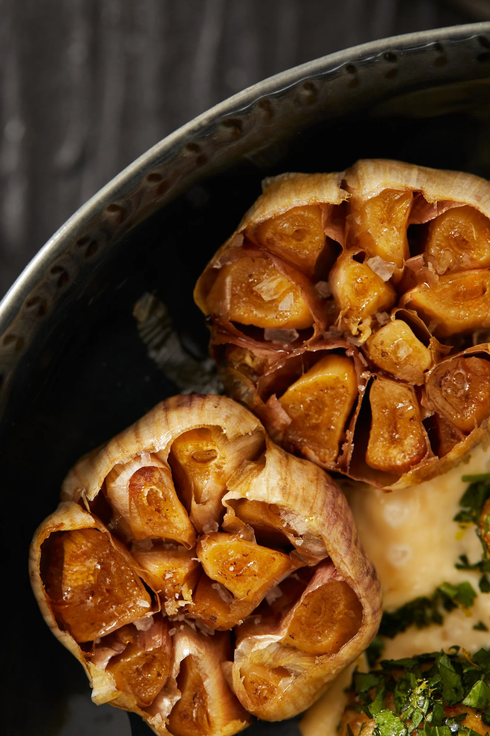 Close-up of roasted garlic bulbs on a dark plate with some parsley and mashed potatoes.