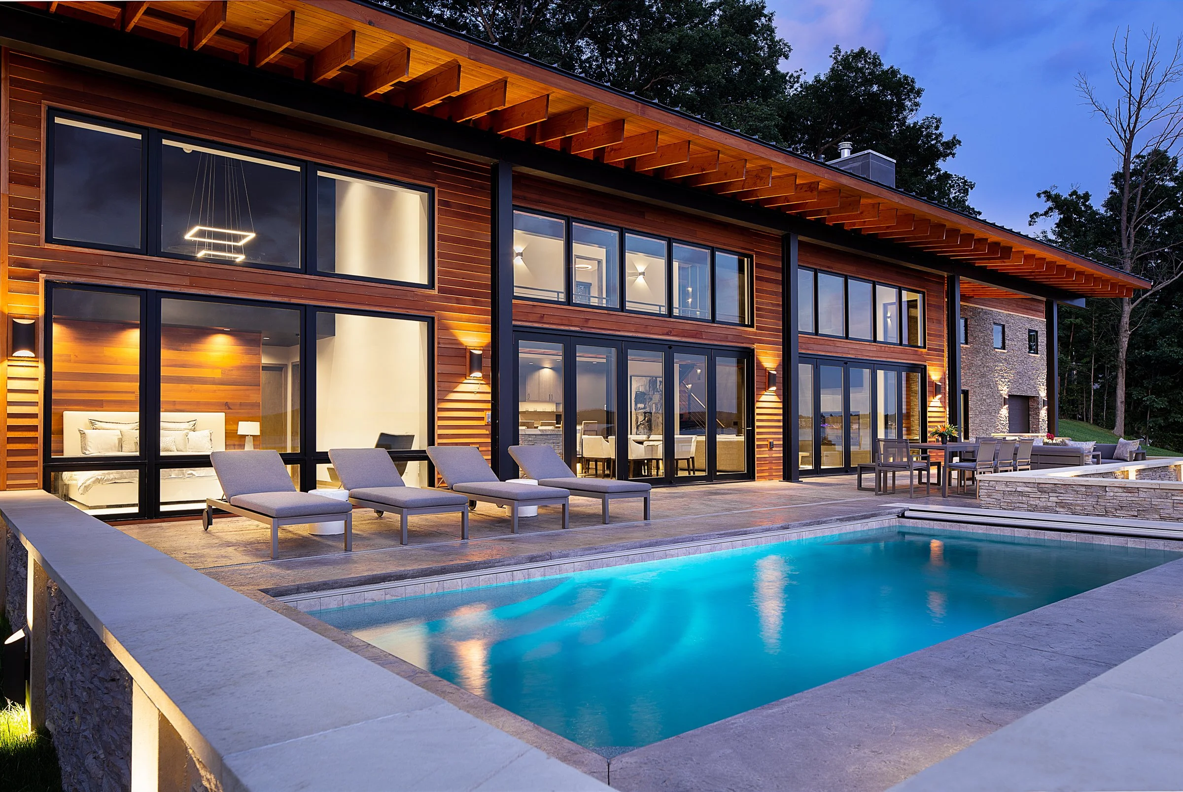 Modern house with large glass windows, outdoor lounging chairs, dining area, and a swimming pool surrounded by stone and wood architecture during evening.