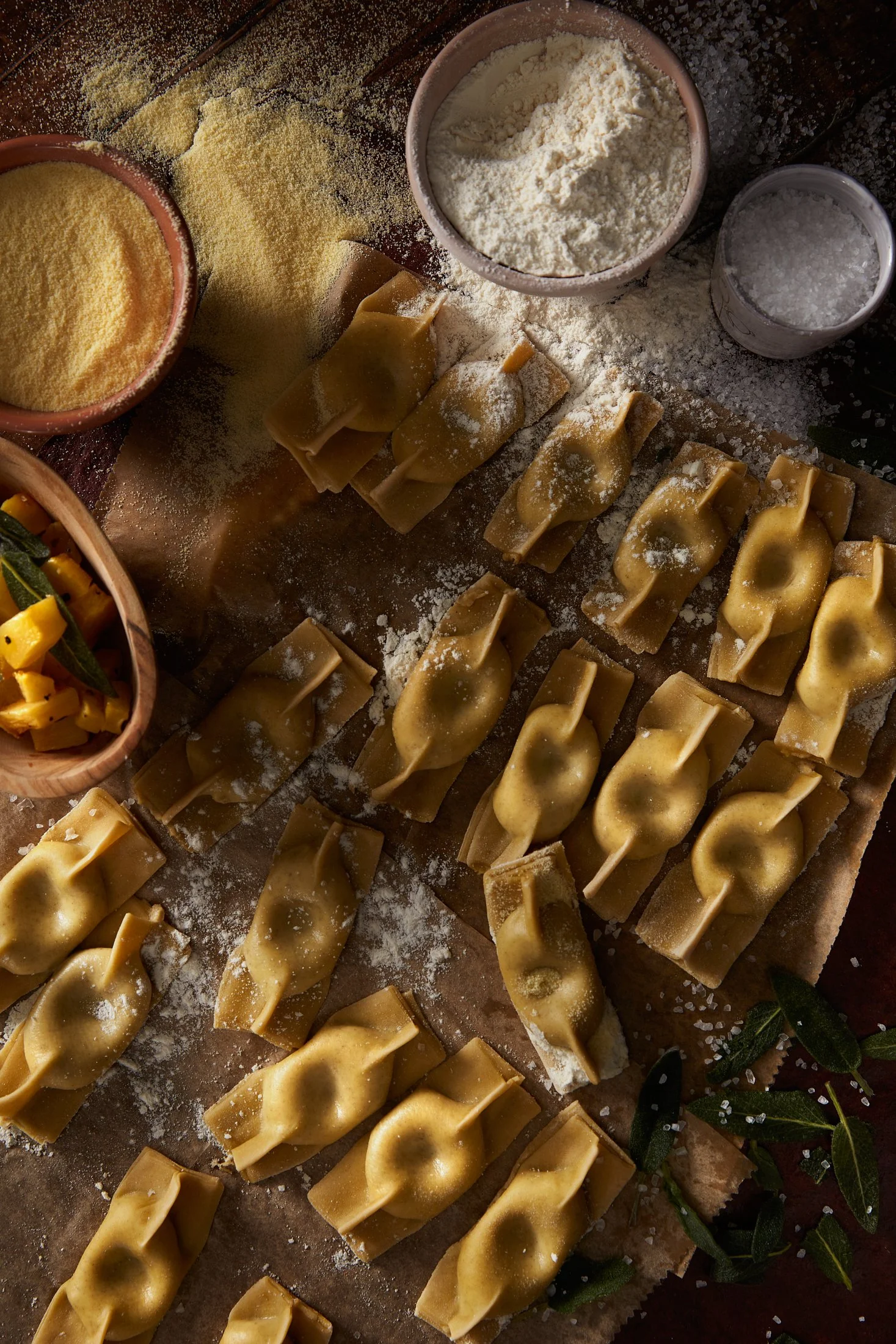 Raw tortellini pasta arranged on a baking sheet with bowls of flour, grated cheese, and salt nearby, along with some fresh sage leaves.