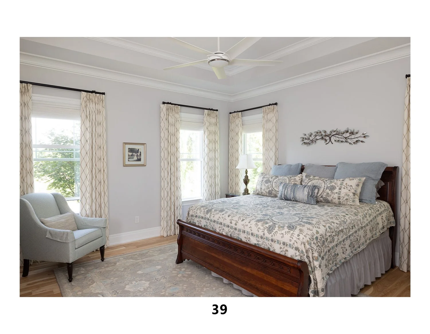 A bedroom with a wooden bed, patterned bedding, pillows, and a wall hanging. There is a nightstand with a lamp, a light armchair with a pillow, three windows with curtains, and a ceiling fan.