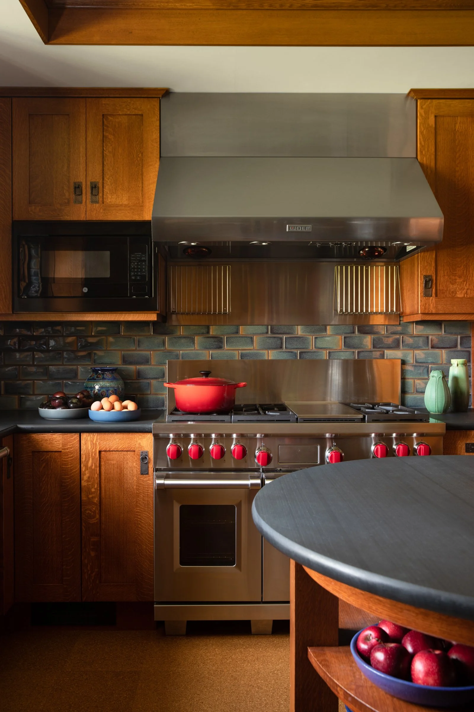 A kitchen with wooden cabinets, stainless steel appliances, and a black countertop; a red pot on a stove, bowls of fruit and eggs on the counter, and decorative vases on the table.