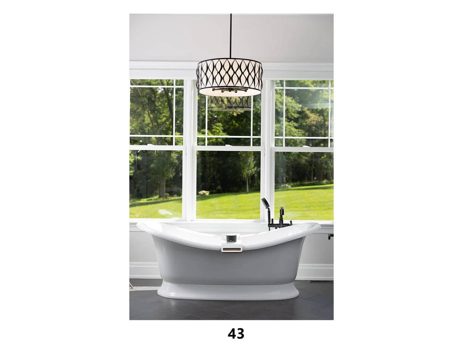A white clawfoot bathtub with black fixtures in front of large window panes showing a green outdoor scene, with a black and white drum pendant light hanging above.