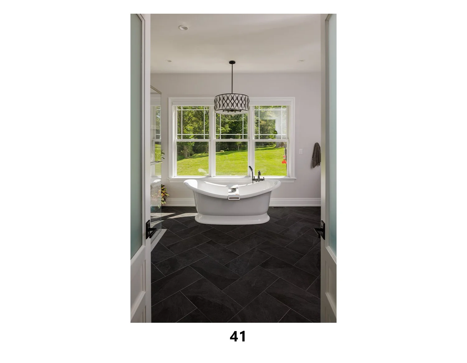 A bathroom with a freestanding bathtub facing a large window showing a green lawn and trees outside. The room has black tiled flooring, white walls, and a black pendant light hanging from the ceiling.