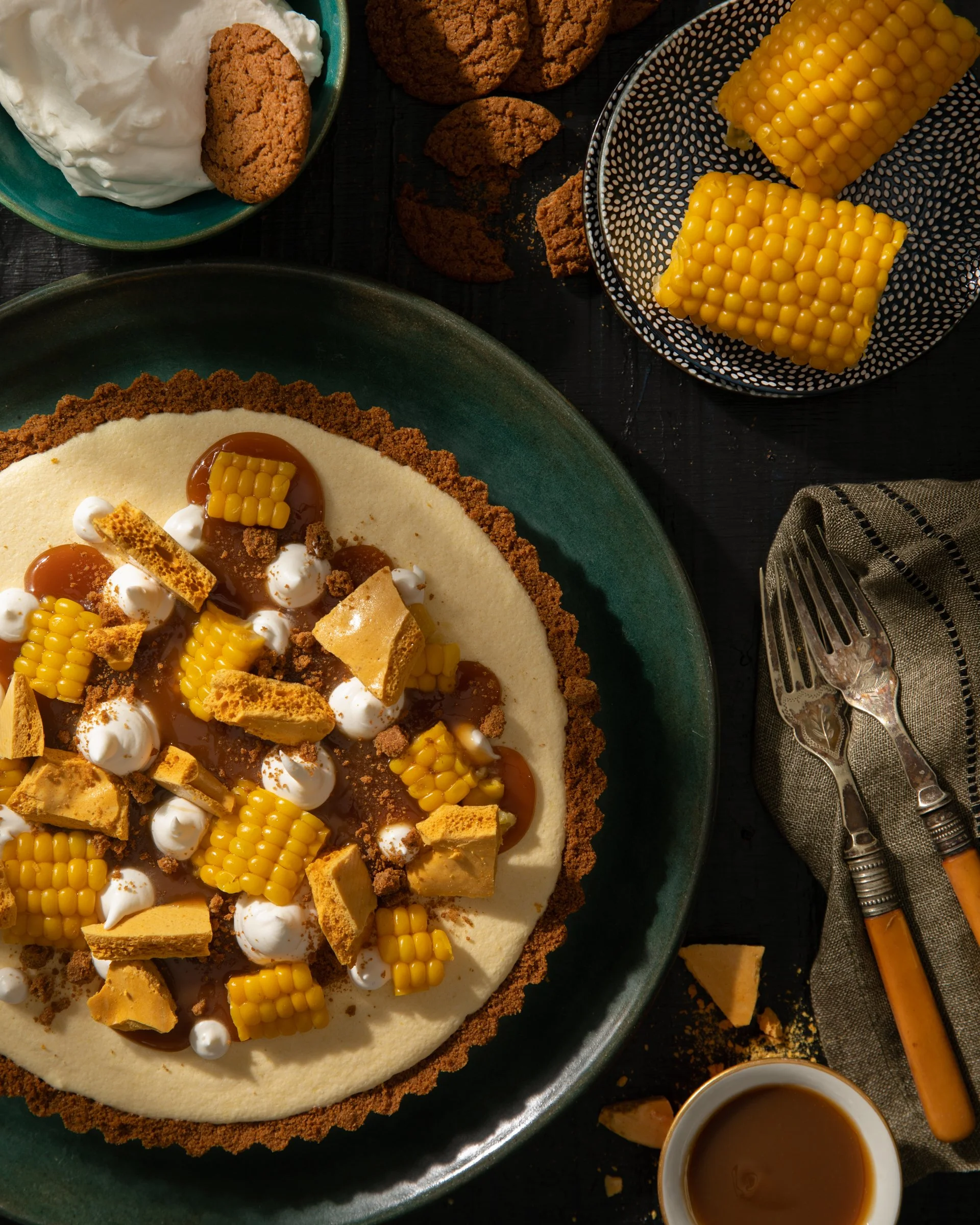 A dessert made with a graham cracker crust, a creamy filling, topped with corn on the cob pieces, dollops of whipped cream, and caramel sauce, served on a green plate with side dishes of cookies and corn, and utensils on a napkin.