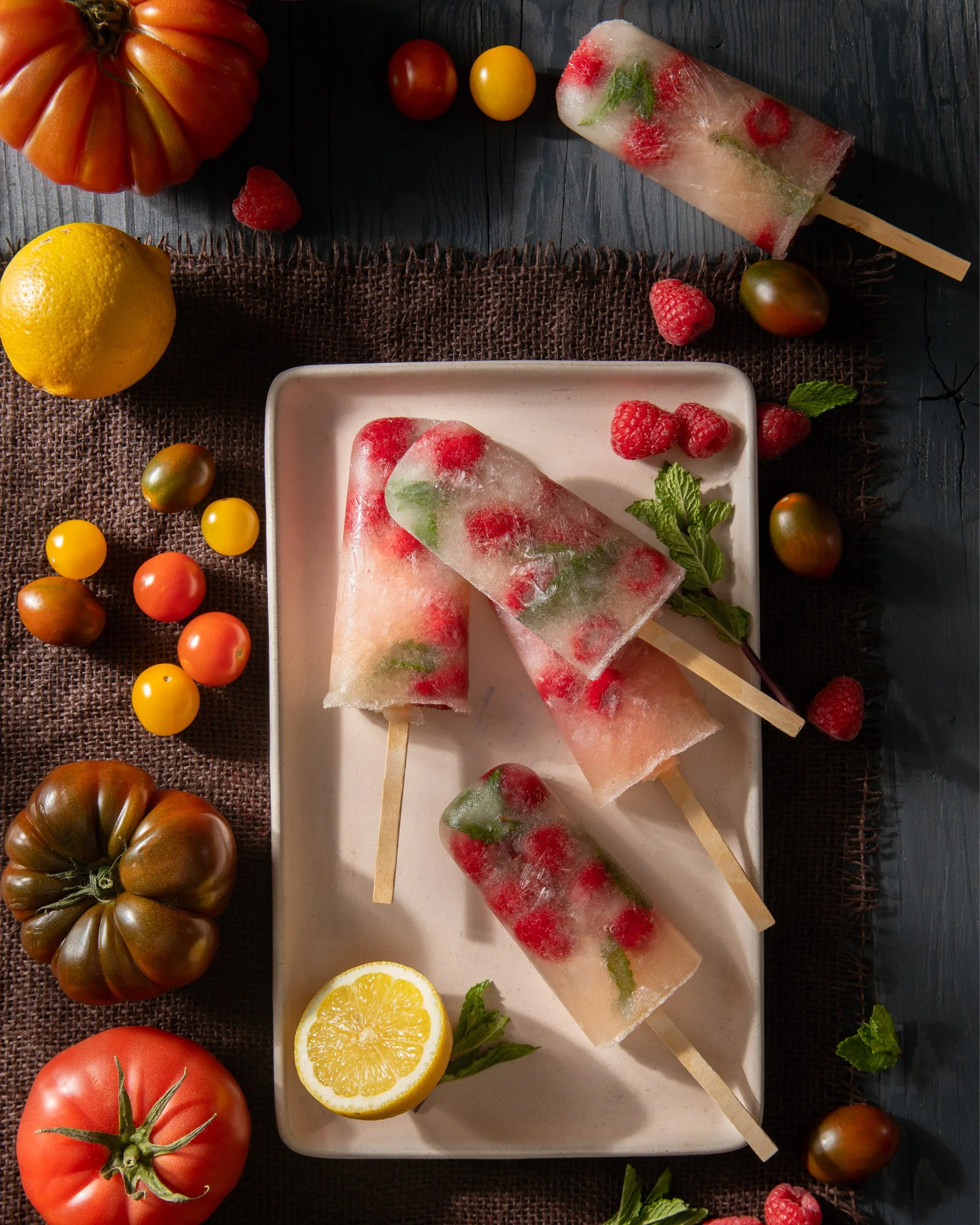 Four strawberry and mint flavored ice pops on a white rectangular plate surrounded by assorted tomatoes, raspberries, lemon, and mint leaves on a dark wooden surface.