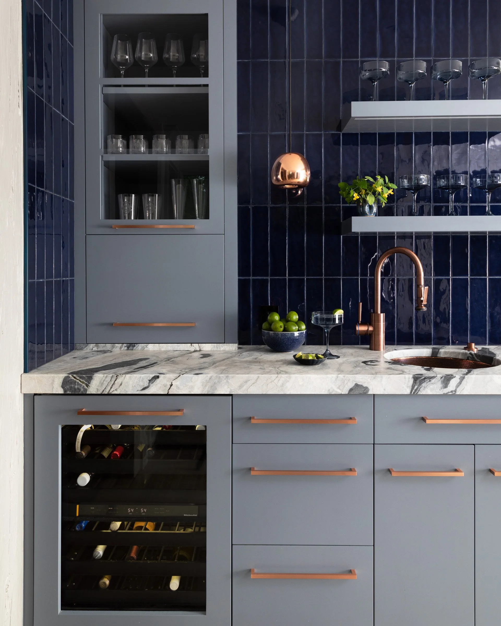Modern kitchen with grey cabinets, marble countertop, navy blue tiled backsplash, and copper fixtures. Glassware is stored on open shelves and inside a glass-front wine cooler. Interior design photography
