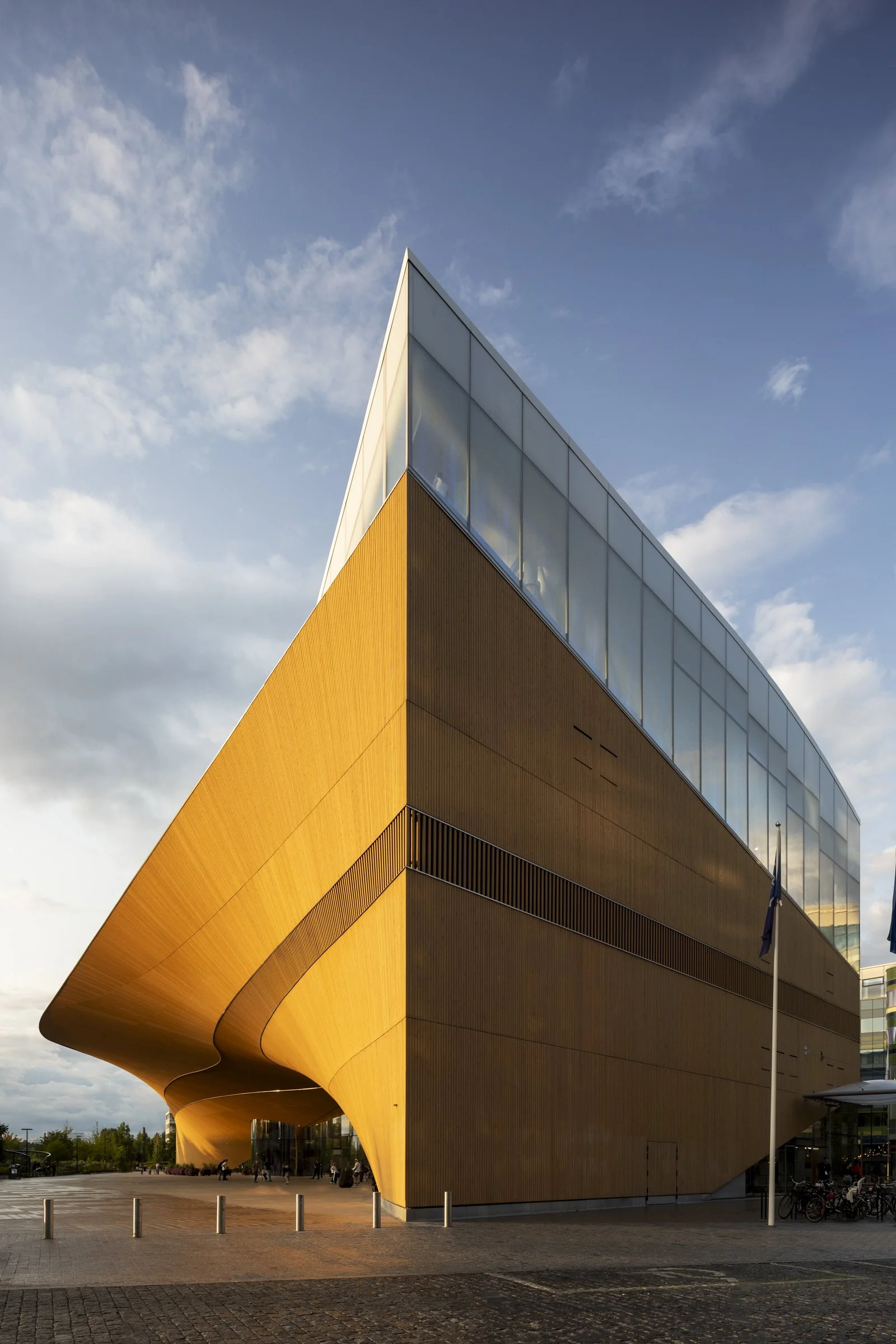 Modern building with a curved wooden exterior and glass top floors, set against a partly cloudy sky.