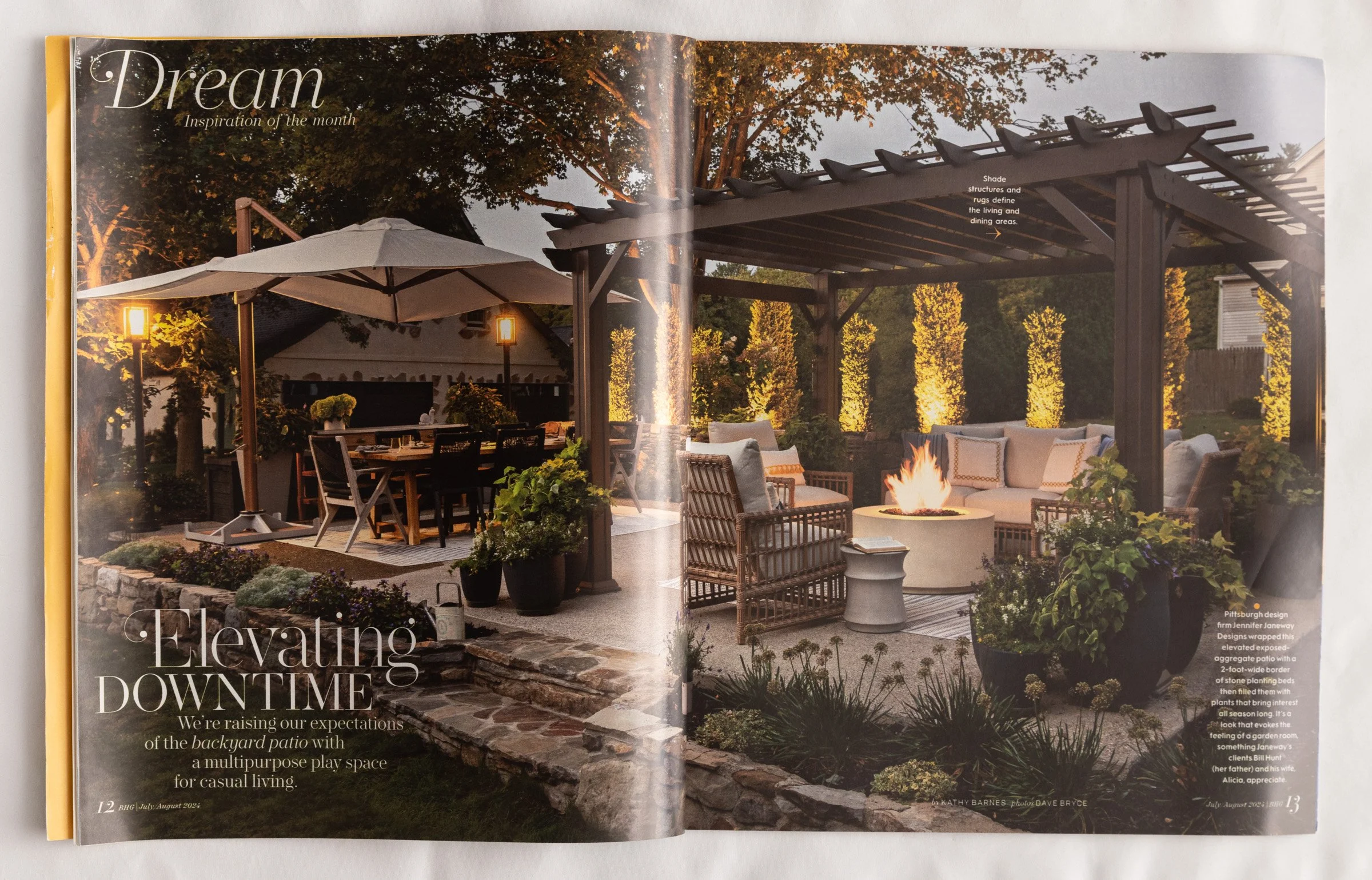A backyard patio with a dining area, a lounge section with a fire pit, and a pergola, illuminated by outdoor lighting, with trees and plants surrounding the space.
