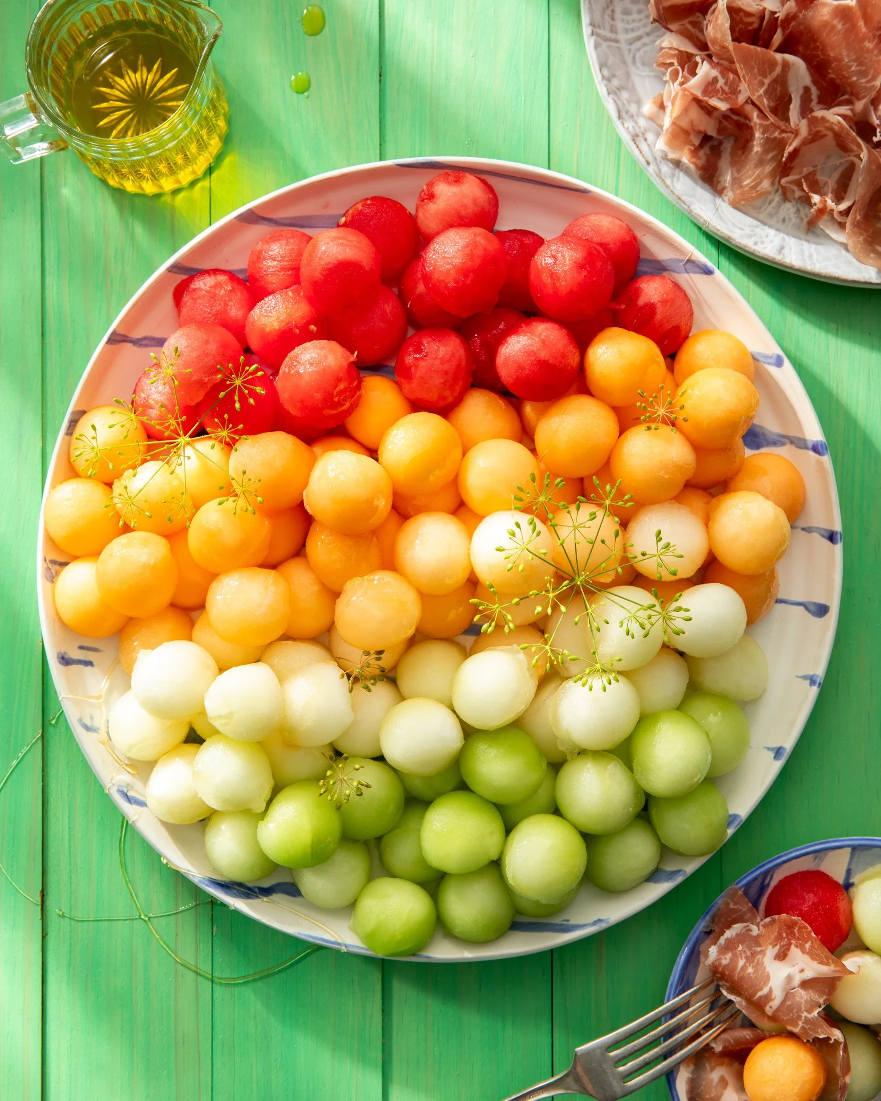 A large plate of assorted colorful melon balls on a green wooden table, with a small bowl of prosciutto, cherry tomatoes, and mozzarella balls, and a small pitcher of olive oil nearby.
