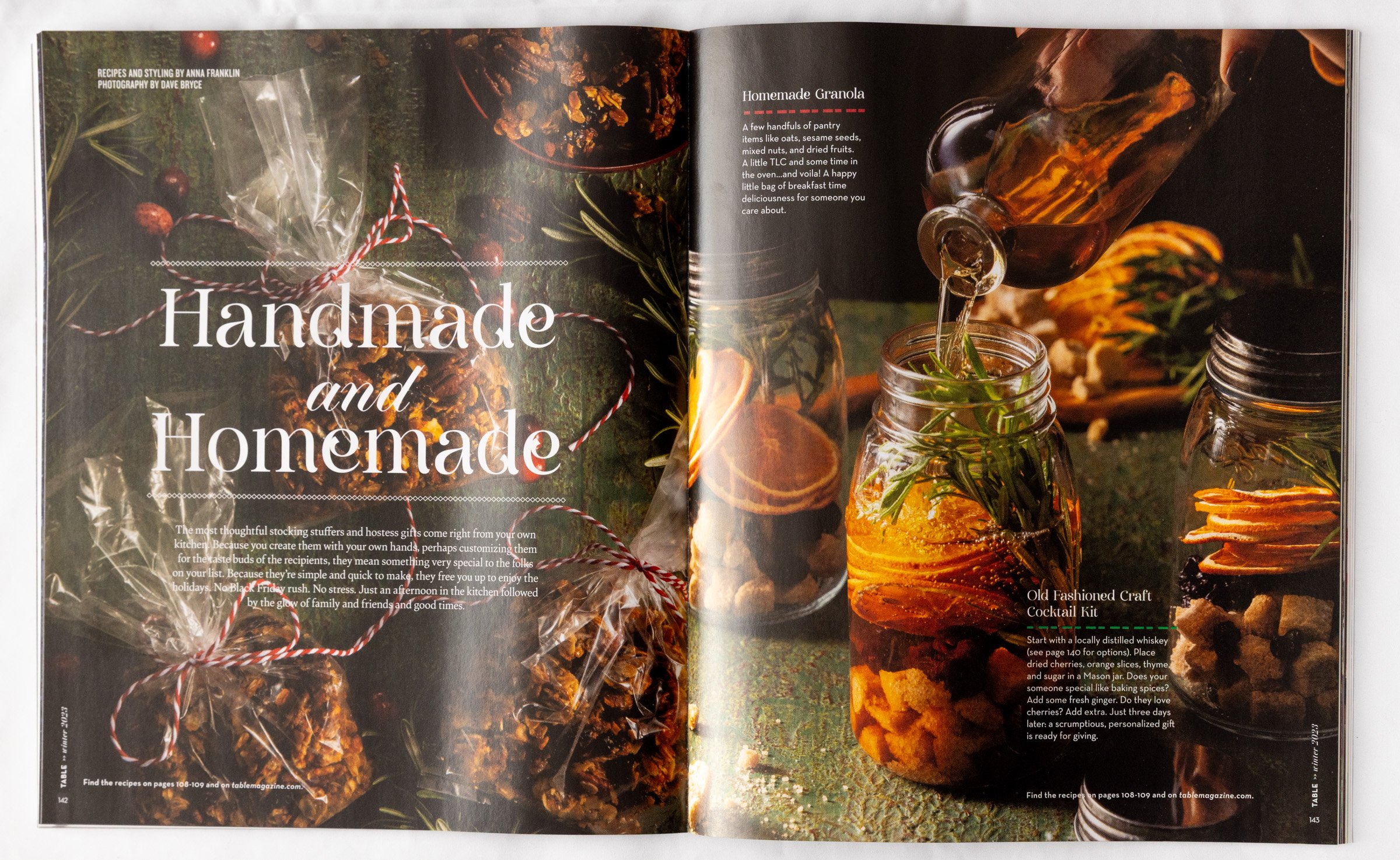 Open magazine spread showing jars of homemade infused spirits with herbs, citrus slices, dried fruits, and spices on a rustic surface, decorated with Christmas ornaments and sprigs of herbs.