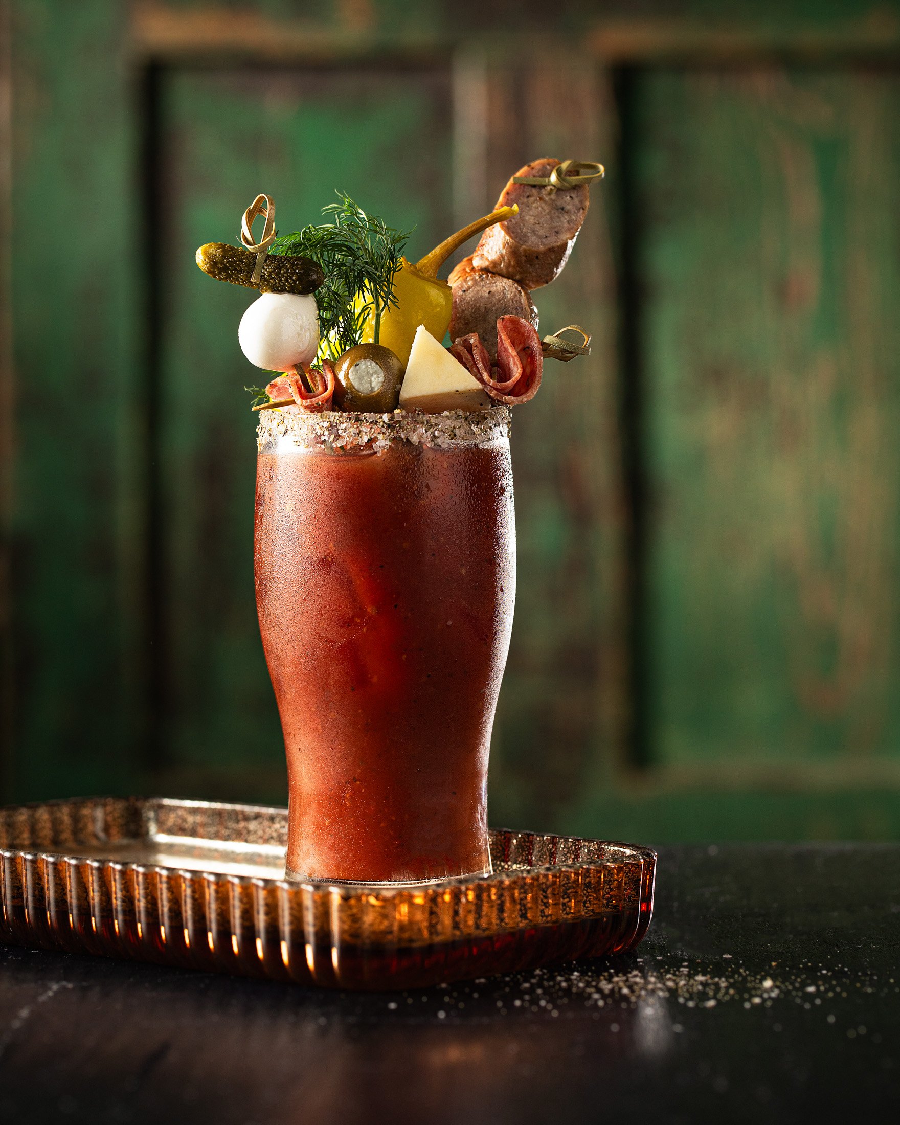 A tall glass of Bloody Mary garnished with olives, pickles, cheese, a lemon, and various meats, placed on a copper tray.
