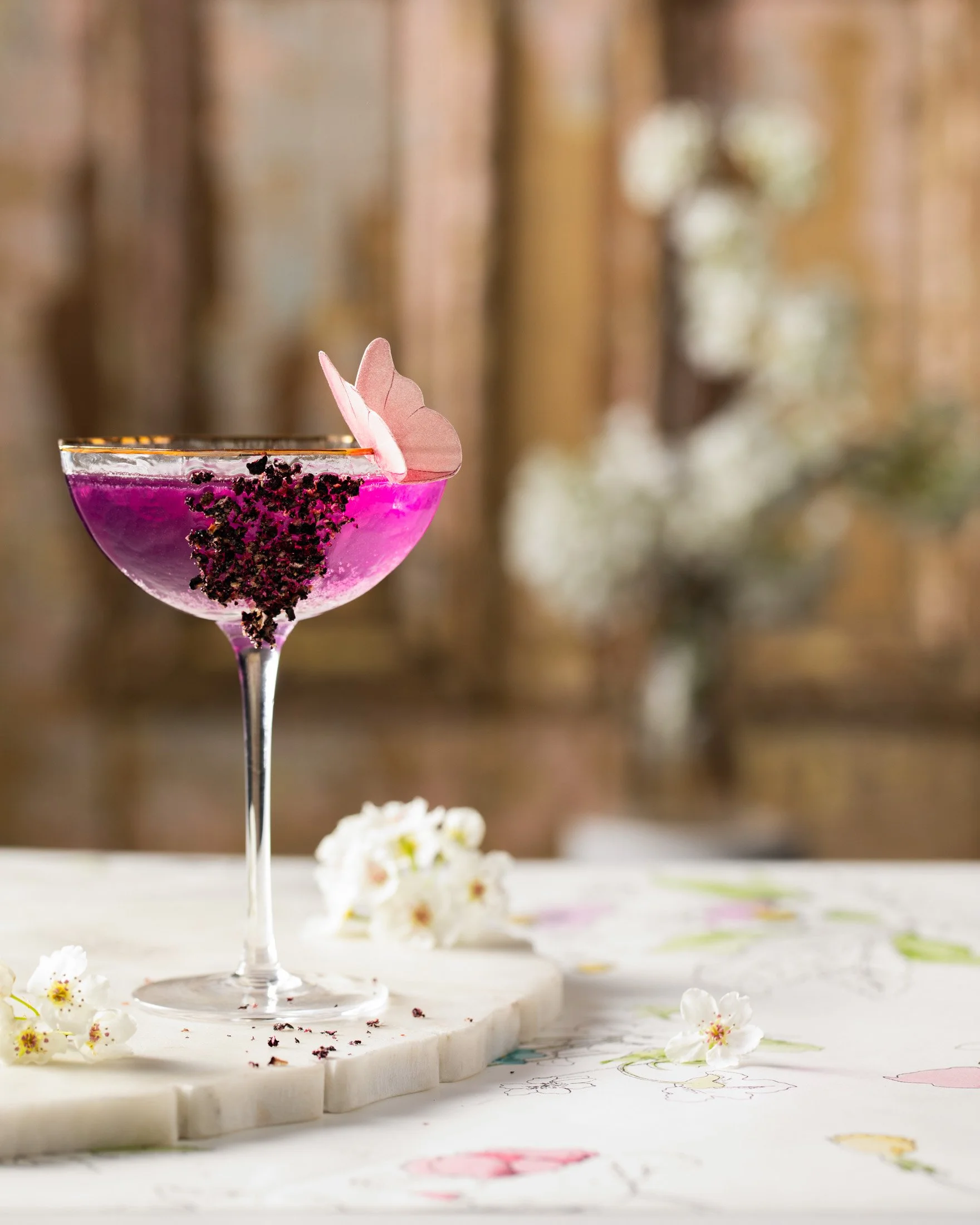 A pink cocktail in a coupe glass garnished with a pink flower petal and blackberry crumbs, on a white marble surface with small white flowers scattered around, with a blurred wooden background and floral decor.