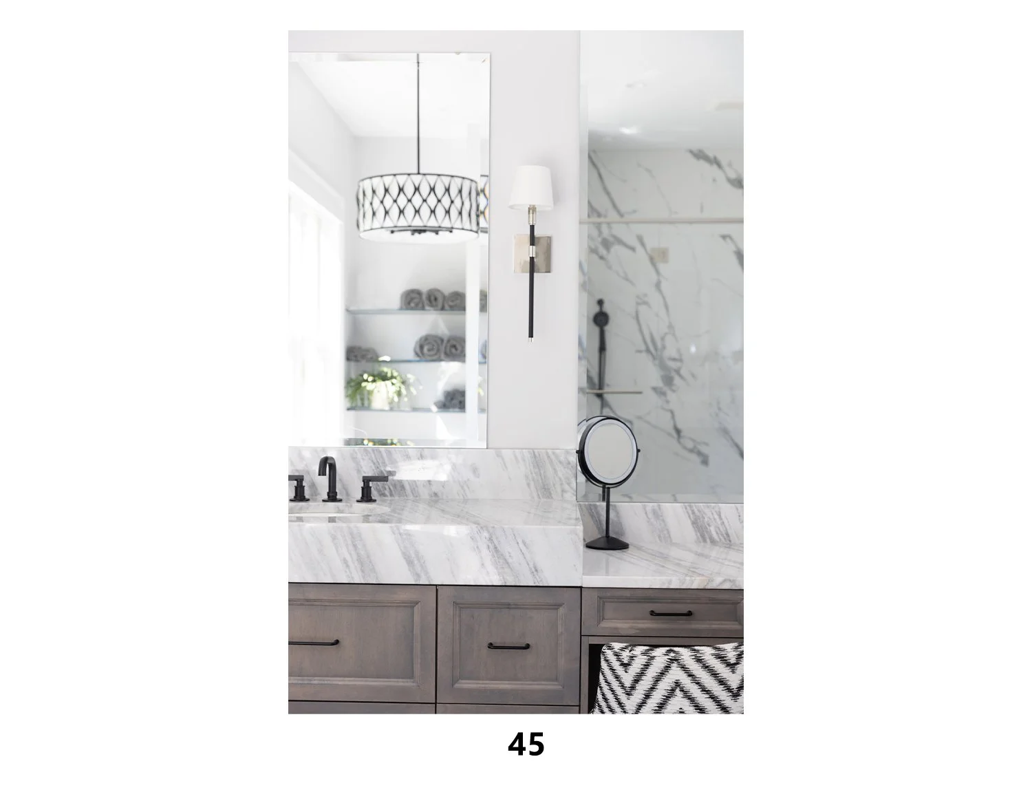 Modern bathroom with marble countertops, a round mirror, black fixtures, a wall-mounted lamp, and shelves with rolled towels and greenery.