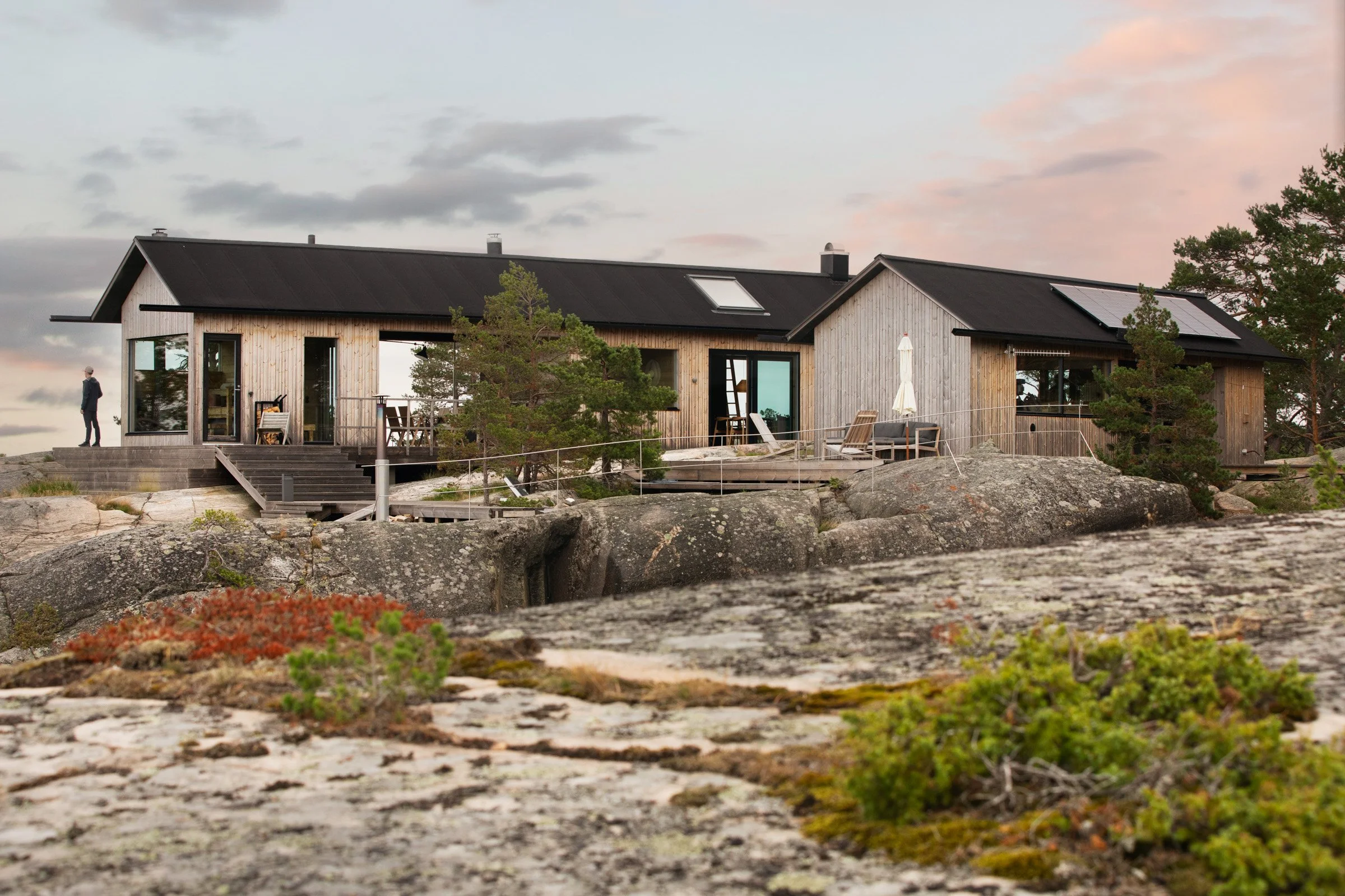 A modern, wooden house on rocky terrain with trees, outdoor furniture, and a person standing on the porch during sunset.