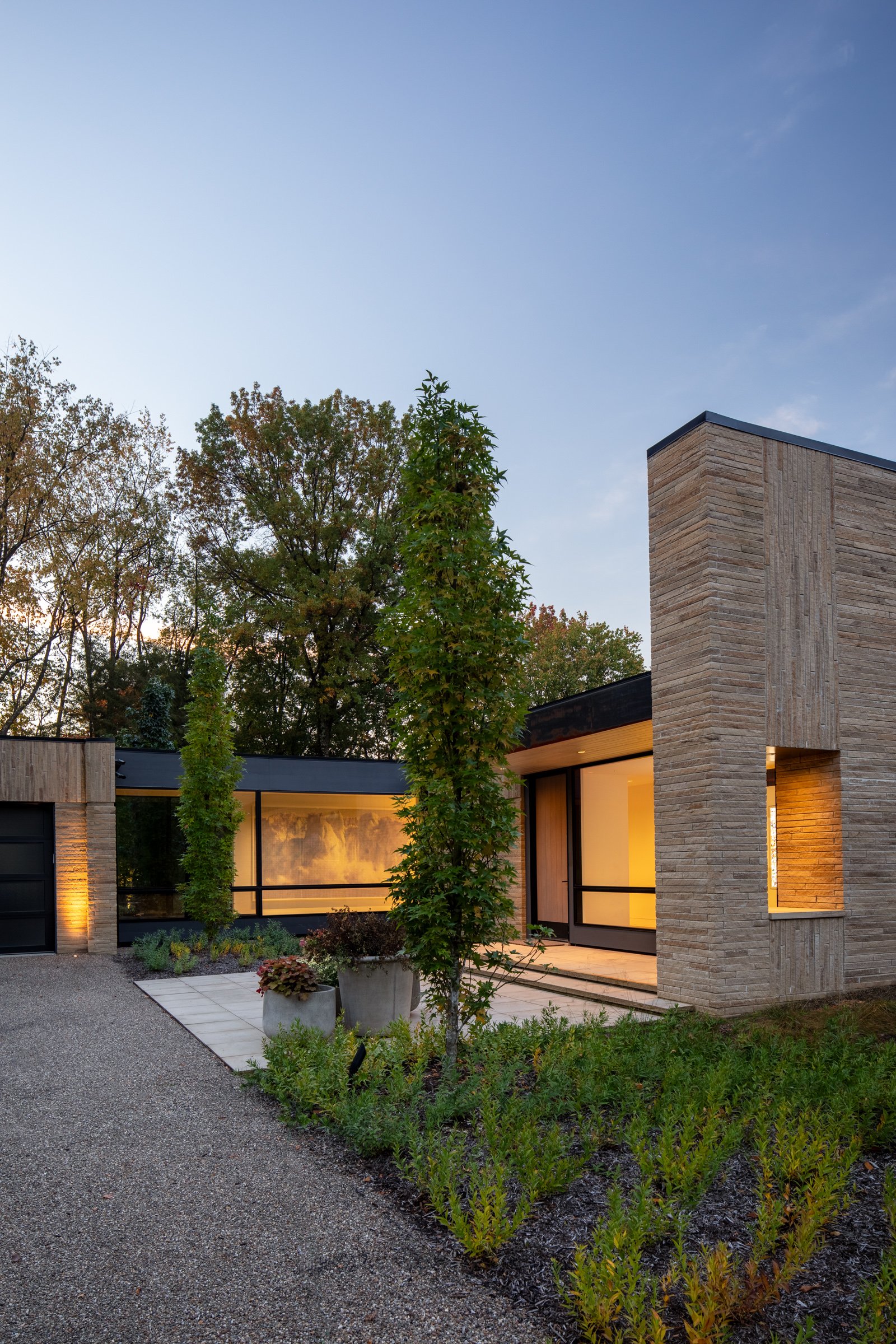 Modern house with large glass windows, stone accents, and landscaped garden with trees and potted plants, sunset lighting.