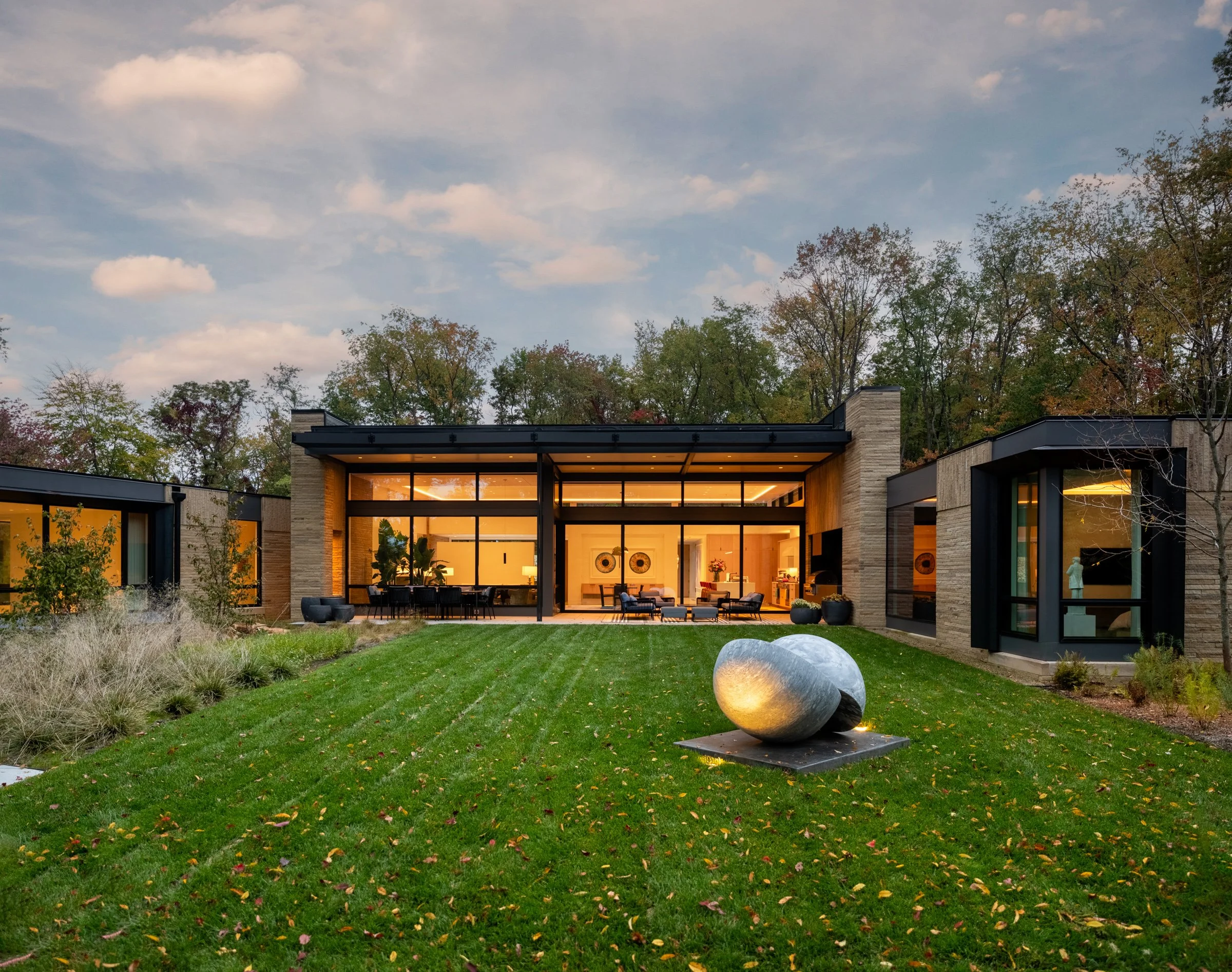 Modern house with large glass windows, landscaped lawn, and abstract stone sculpture outside, surrounded by trees at dusk.