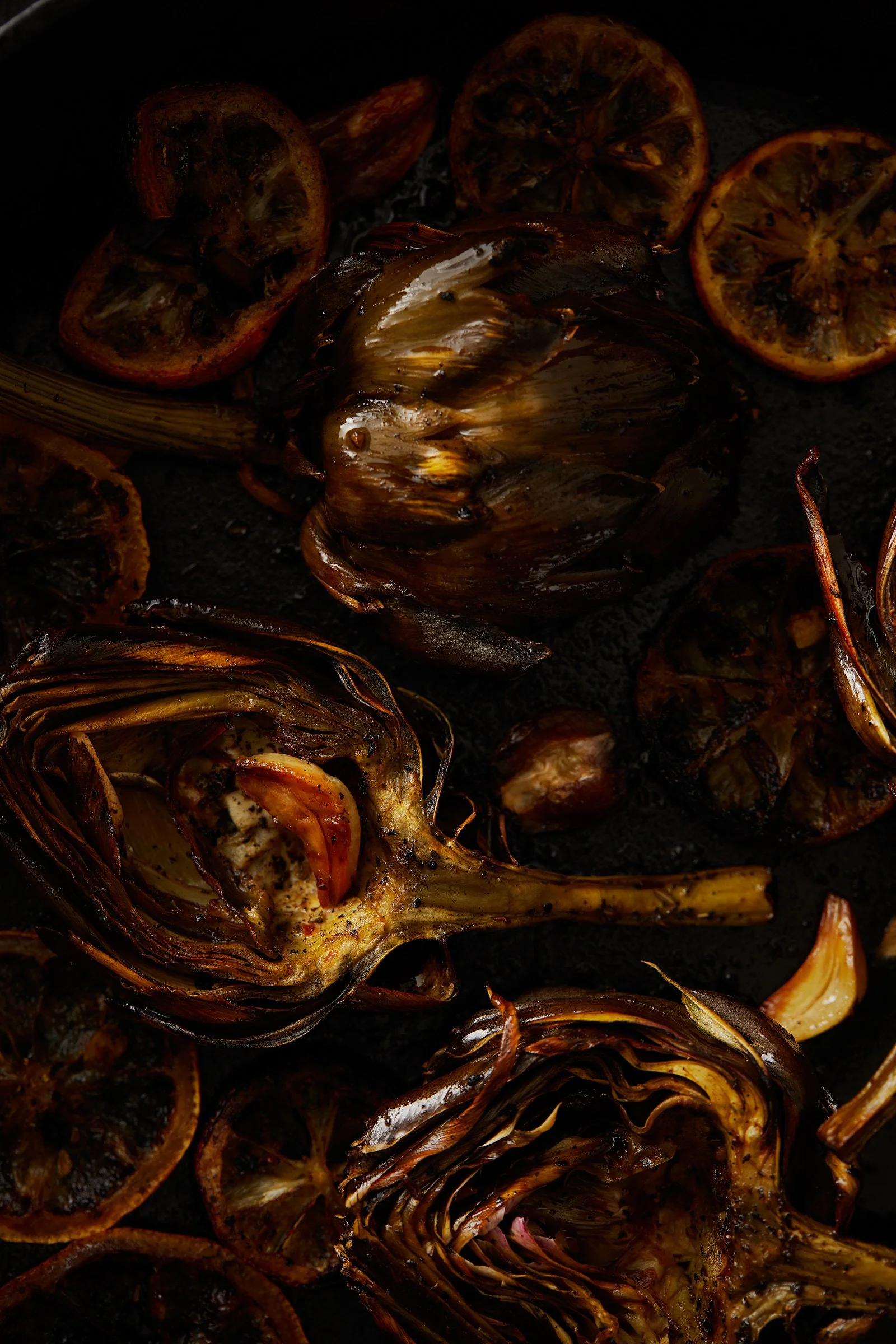 Charred artichokes and lemon slices on a dark surface, with some garlic cloves.