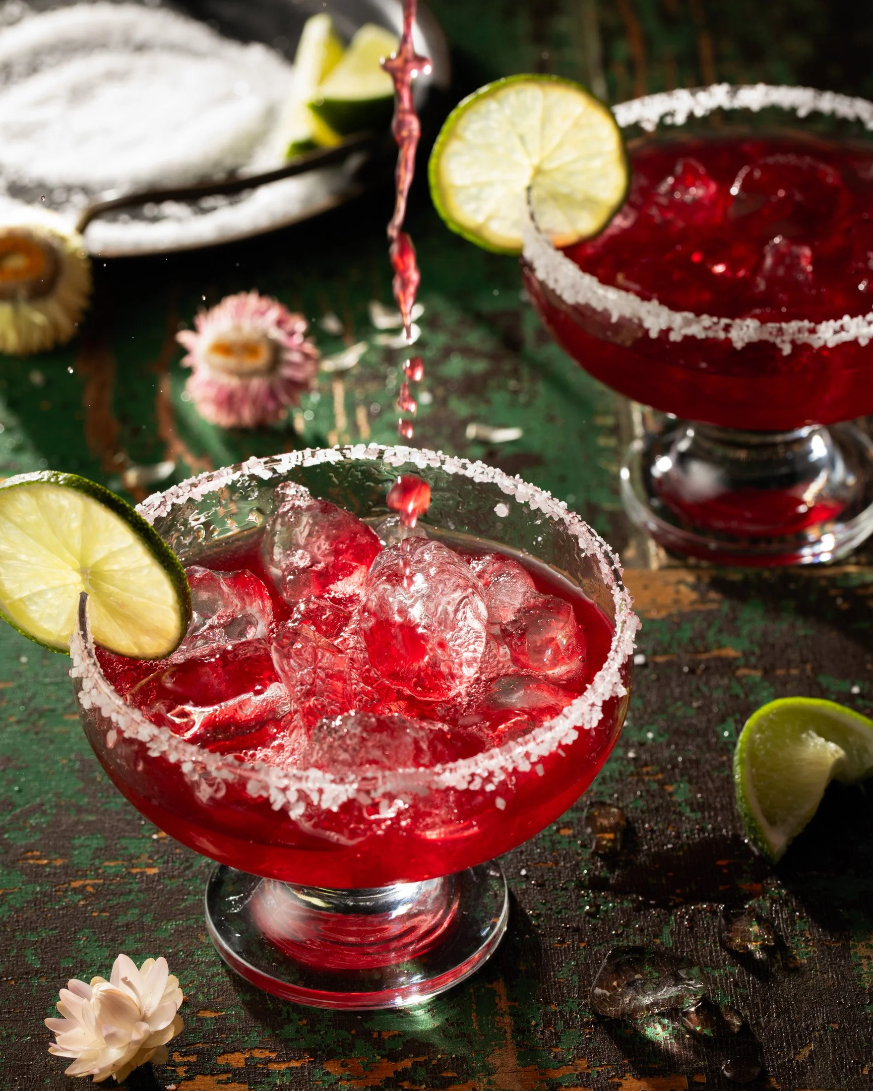 Two glasses of red cocktail with salted rims and lime wedges, one glass being filled with a splash of lime juice, on a dark textured surface with lime slices, flowers, and spilled ice.