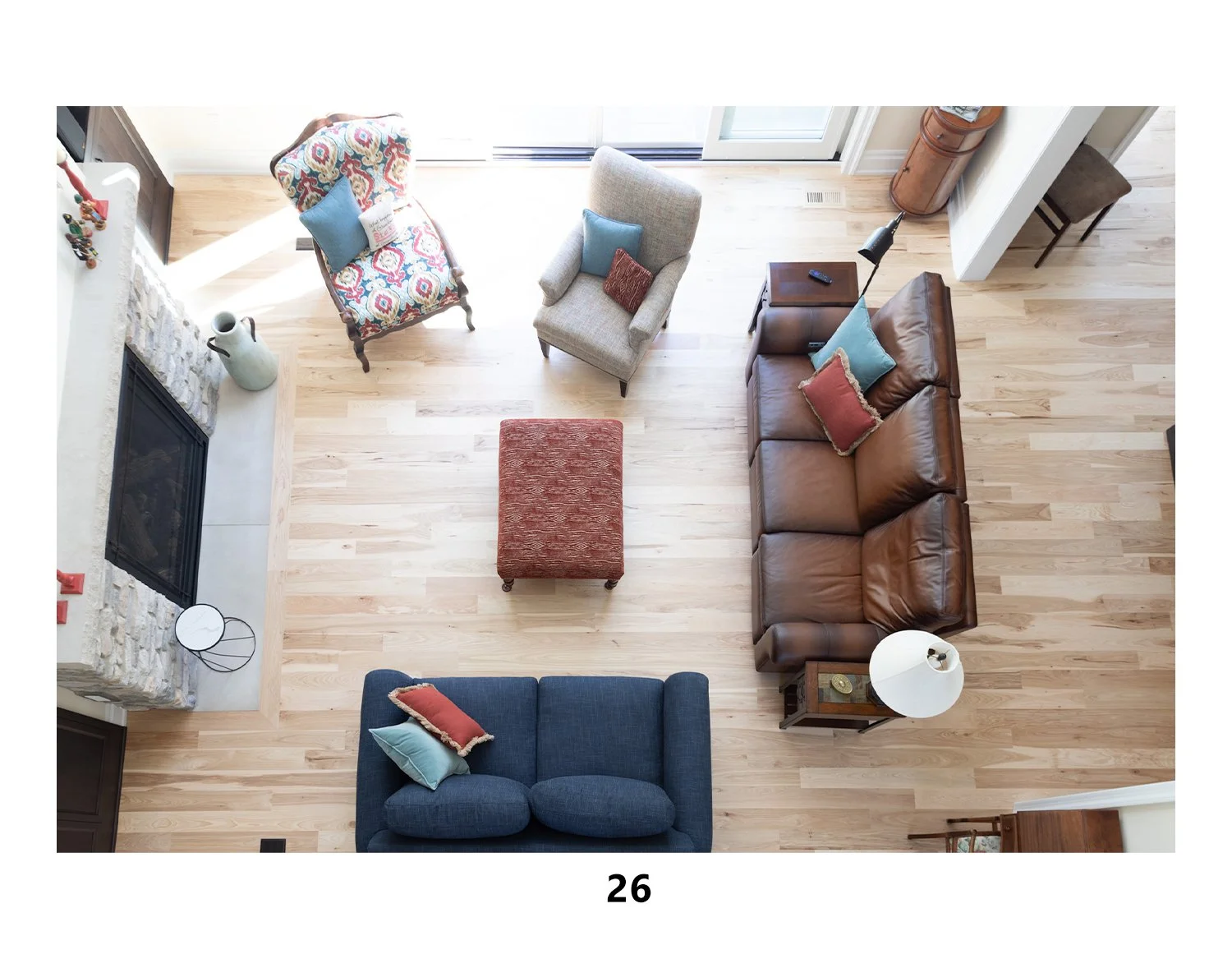 Top-down view of a living room with a fireplace, three couches (blue, beige, brown), an orange ottoman, a small side table with a lamp, three cushions on the couches, and a closed door leading outside.