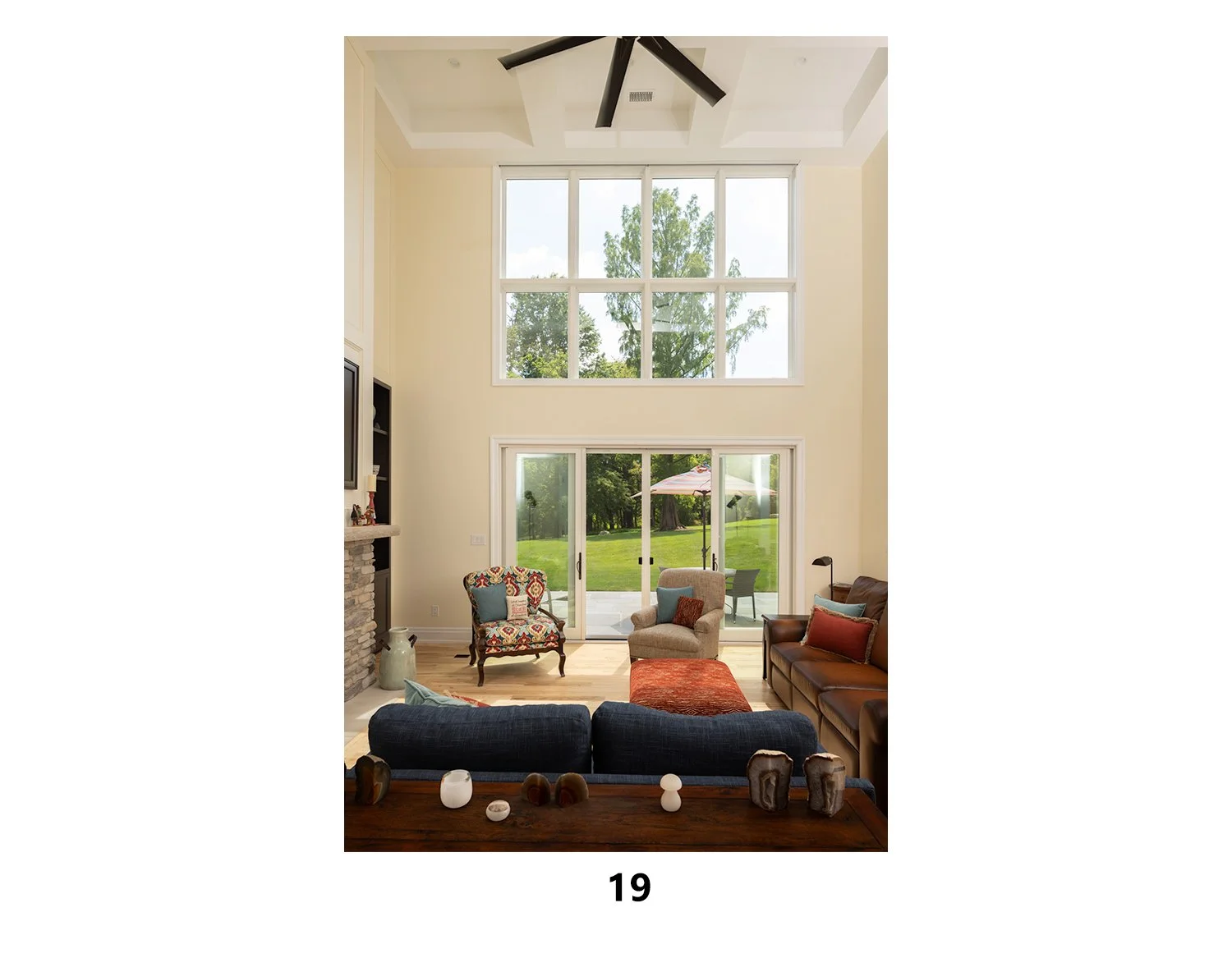 Living room with large windows and sliding glass doors looking out to a green backyard with patio furniture and an umbrella. The room has a high ceiling with a fan, a stone fireplace, and comfortable chairs and sofa.