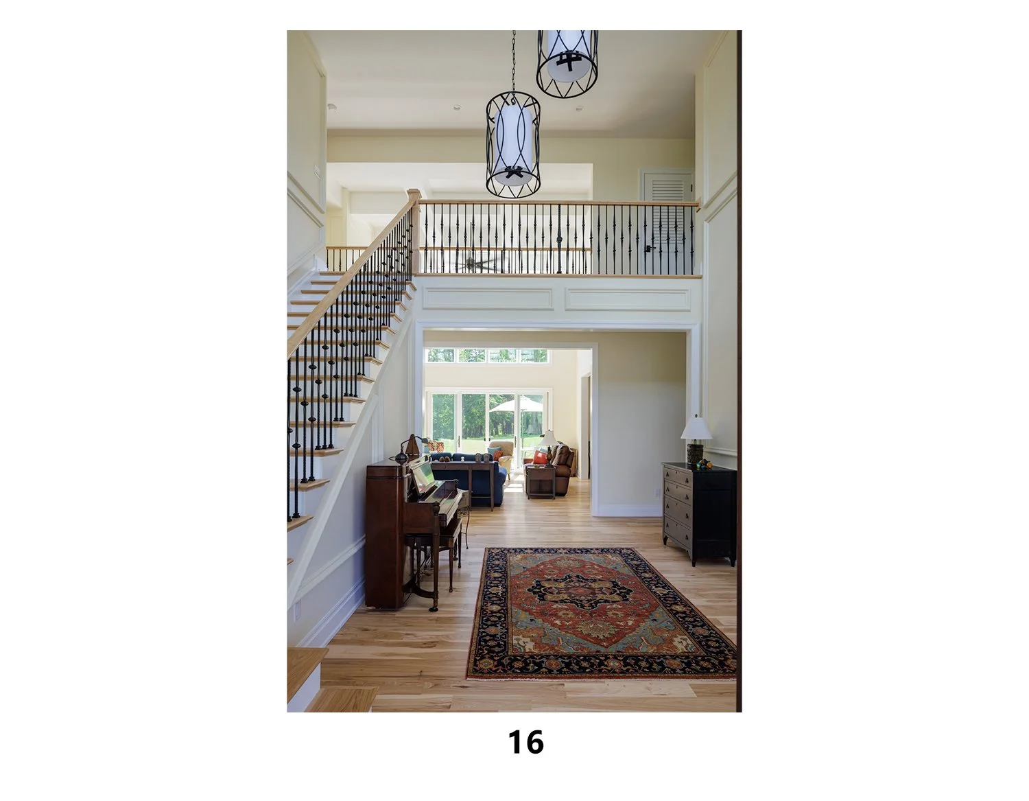 Interior view of a two-story house with a staircase leading to a second-floor balcony overlooking a living room. The living room has large windows with greenery outside, a piano, a rug, and various seating and furniture.