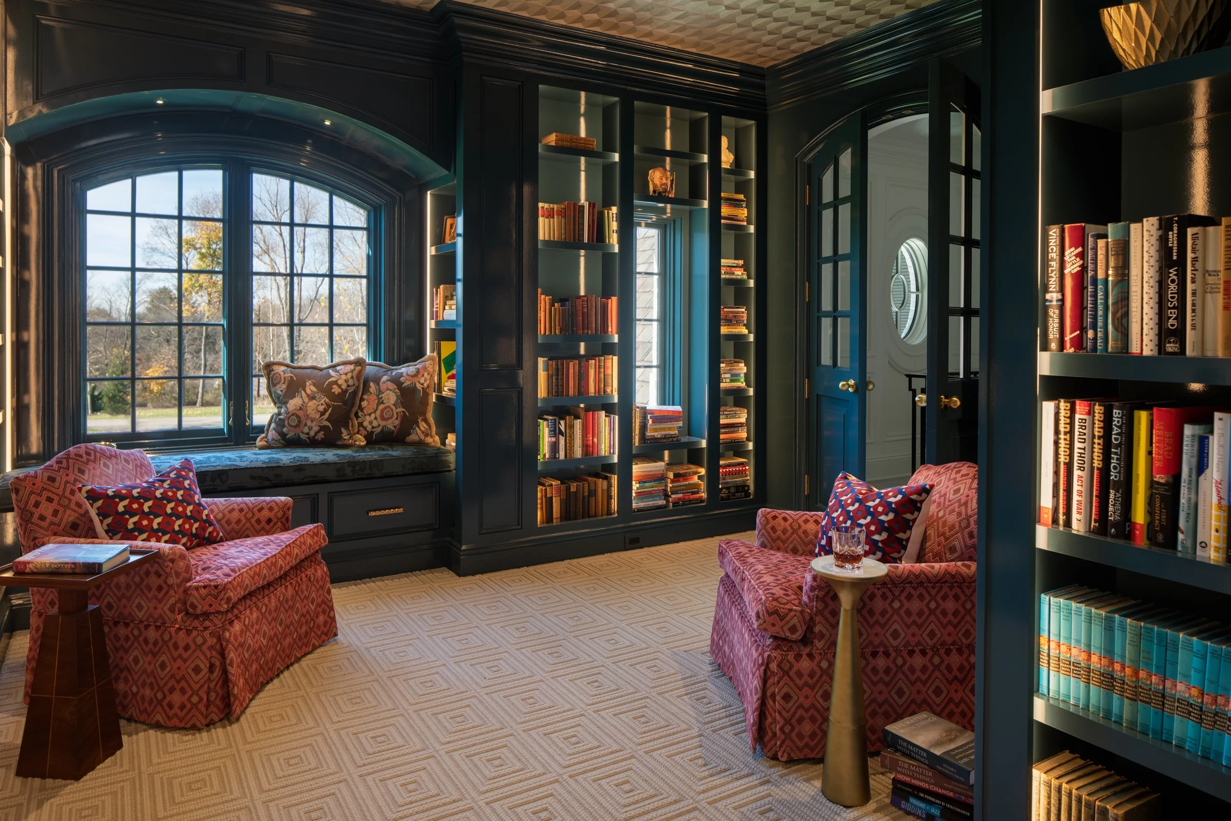 A cozy library room with blue bookshelves filled with books, a window seat with cushions, two pink patterned armchairs with cushions, a side table with a glass of whiskey, and a beige carpet with a diamond pattern.