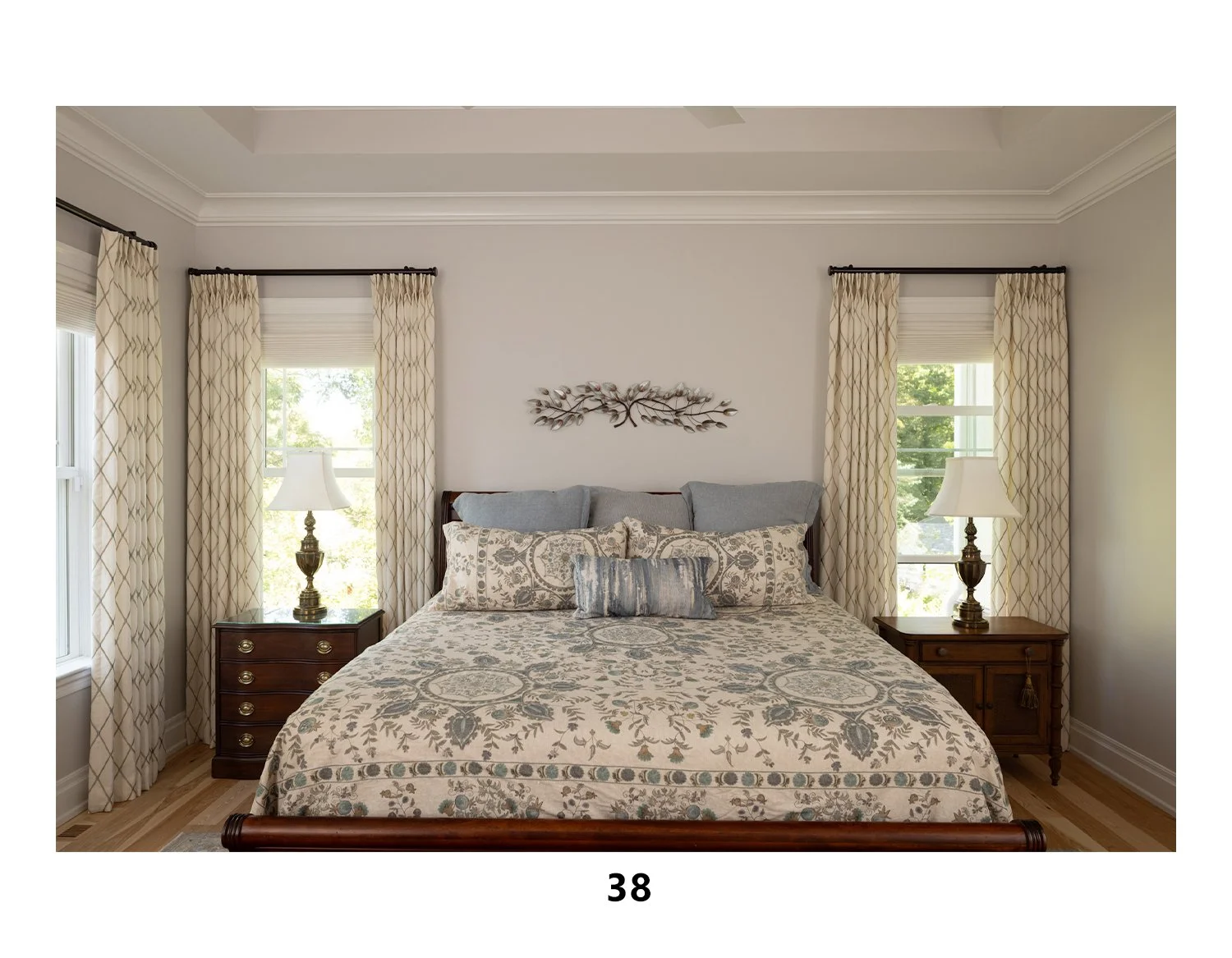 A bedroom with a bed, patterned bedding, and pillows, flanked by two nightstands with lamps, curtains on windows, and a decorative wall hanging above the bed.