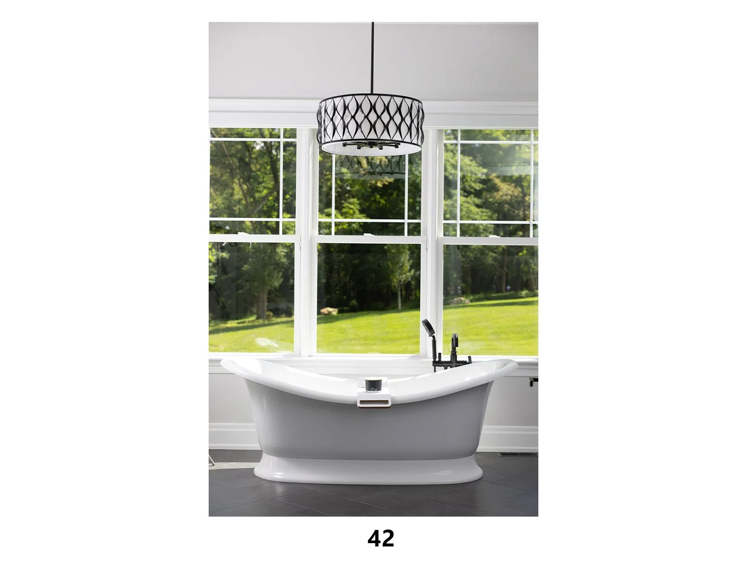 A white bathtub with a black faucet sits in front of large windows showing green trees and grass outside. A black pendant light hangs from the ceiling.