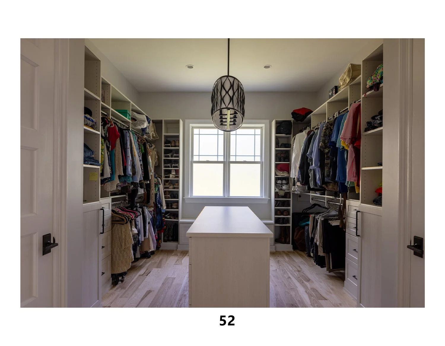 A walk-in closet with white shelves filled with folded clothes, shoes, and accessories. There is a large window in the center, and a white island in the middle of the room. A hanging light fixture is above the island.