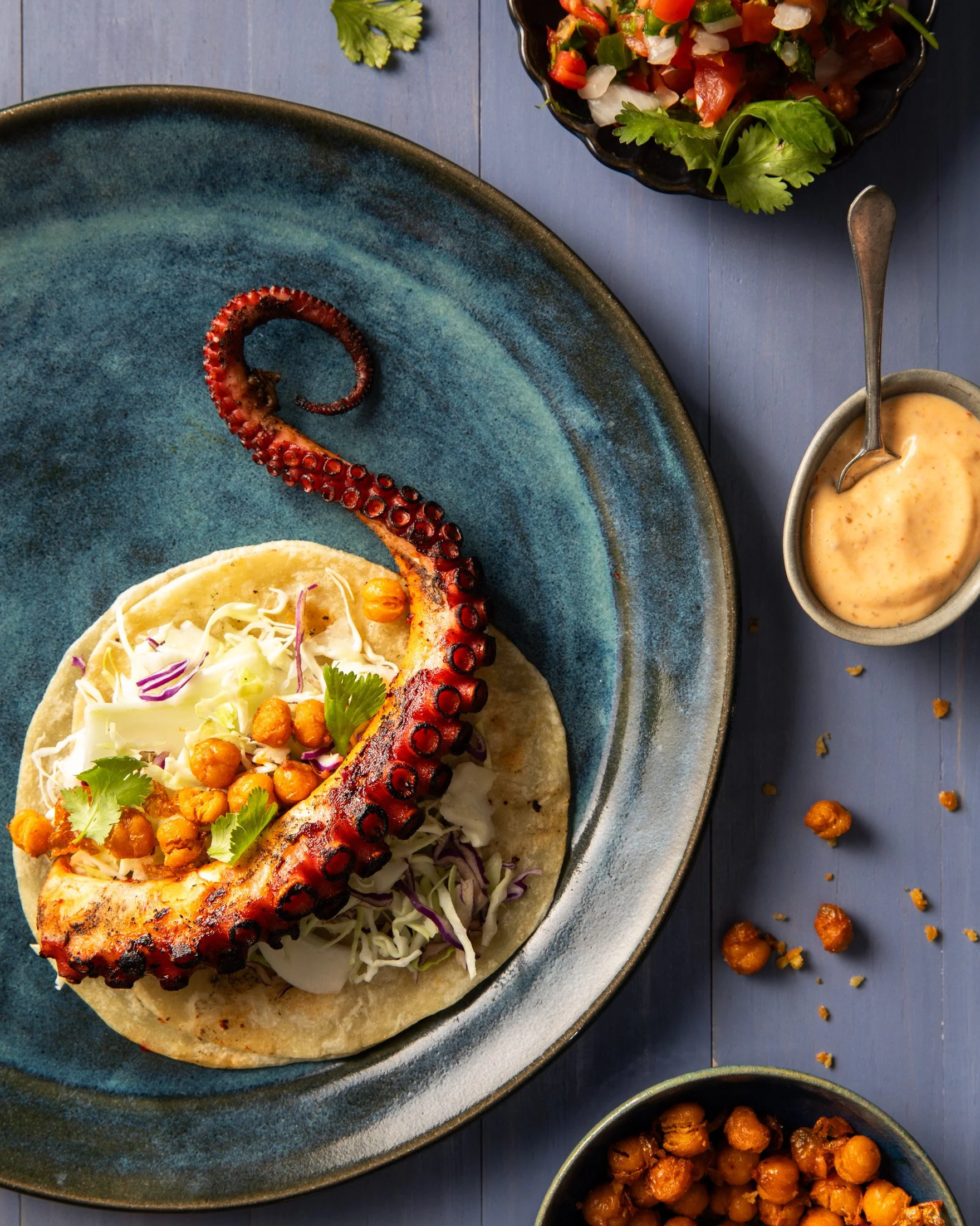 Grilled octopus tentacle on a taco with cabbage, chickpeas, cilantro, in a soft tortilla, served on a blue plate with side salads and sauces.