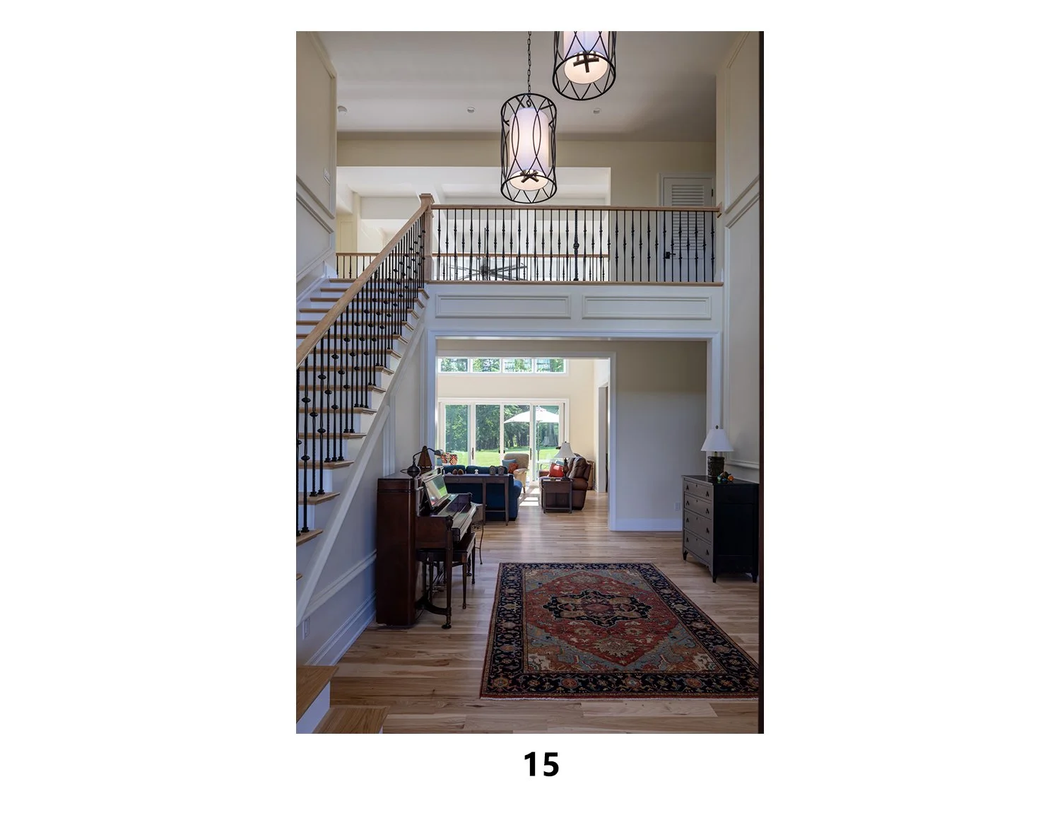 Interior view of a spacious living area with a staircase on the left, a mezzanine with a railing, and a door leading to a sitting room with large windows and outdoor view. The room has hardwood floors, a patterned area rug, and scattered furniture in