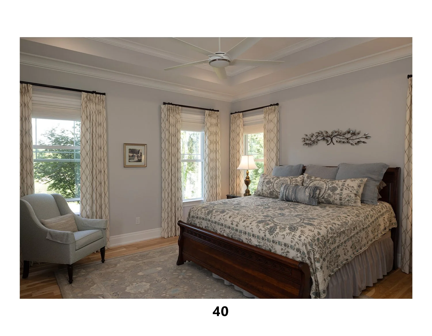 A bedroom with a wooden bed, patterned bedding, multiple pillows, a bedside lamp, a small armchair, and windows with cream-colored curtains.