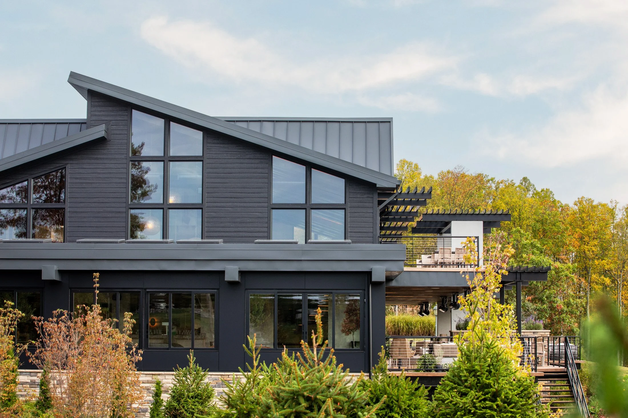 Modern two-story house with dark exterior and large windows, surrounded by greenery and trees.