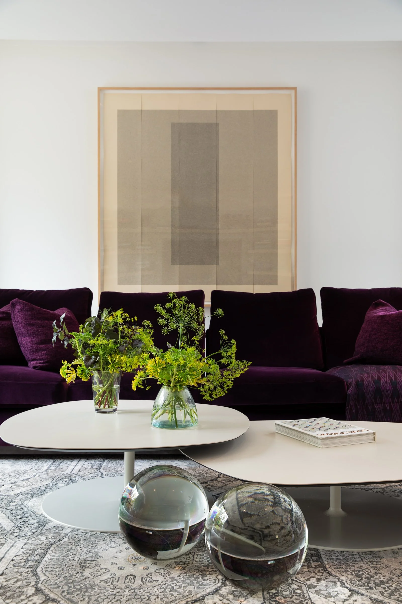 Modern living room with white oval coffee tables, purple velvet sofa with matching pillows, two glass vases with green plants, abstract art on white wall, patterned rug, and glass sphere decor.