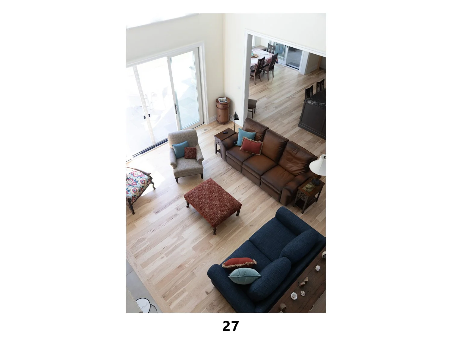 Living room with a brown leather sofa, blue armchair, red ottoman, and a blue sofa, with a sliding glass door, wooden flooring, side tables, and cushions.