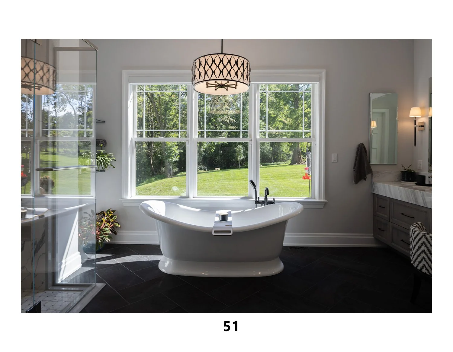 Modern bathroom with a freestanding bathtub, large window showing a green backyard, vanity with mirror, plant, and a shower area.