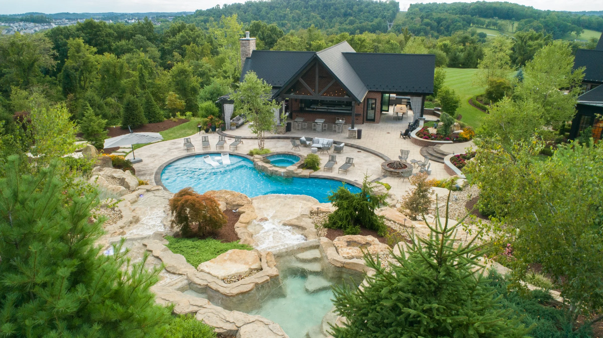 A backyard with a swimming pool, hot tub, and a waterfall feature, surrounded by trees and landscaped garden beds. There is a patio area with chairs, a fire pit, and an outdoor kitchen or bar, with a house in the background.