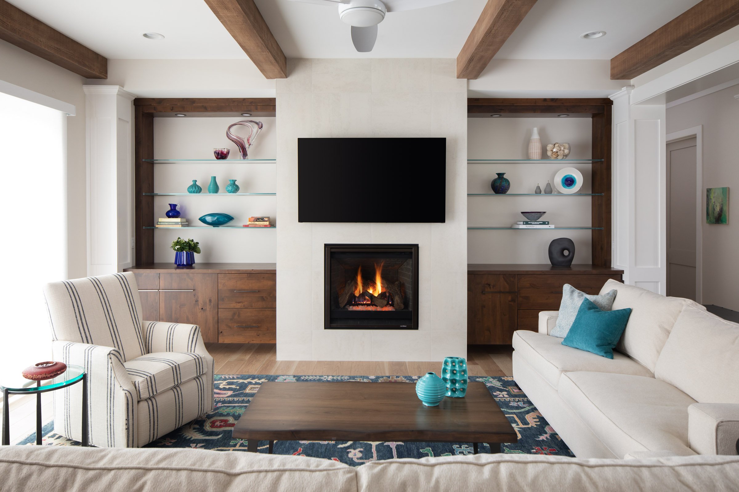 Living room with fireplace, flat-screen TV above it, built-in shelves with decorative items on both sides, beige sofa with blue and gray pillows, striped armchair, coffee table with blue vases, patterned rug, and wood ceiling beams.