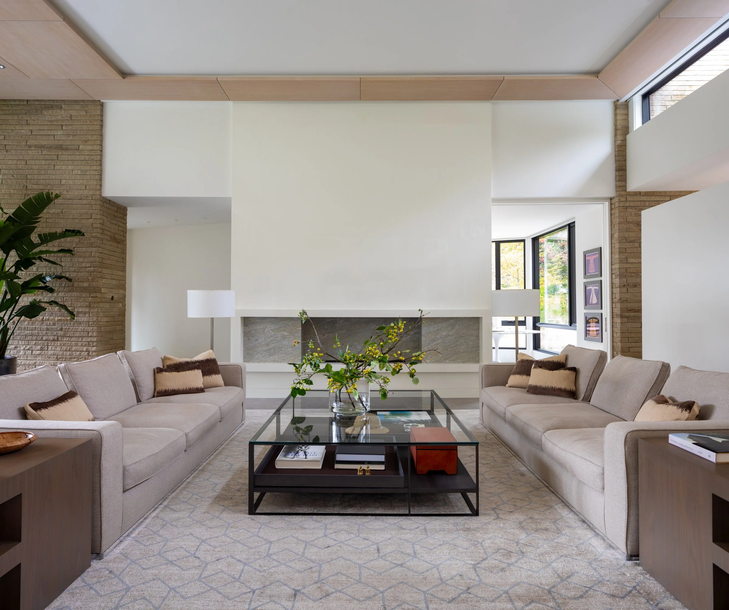 Modern living room with two beige sofas, a glass coffee table with a flower arrangement, and large windows with black frames.