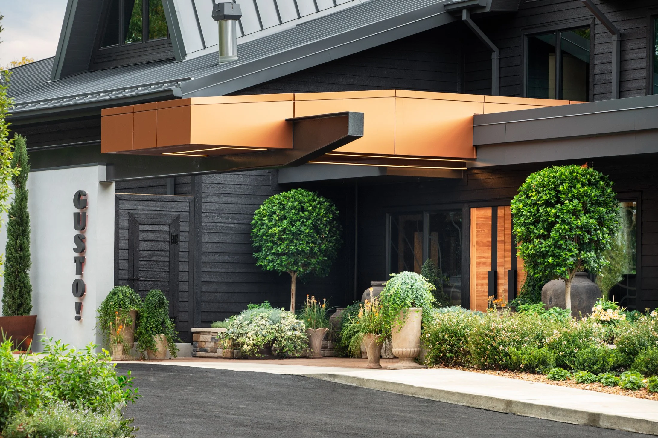 Modern building entrance with black wooden siding, bronze-colored awning, and lush green plants and trees in decorative pots.