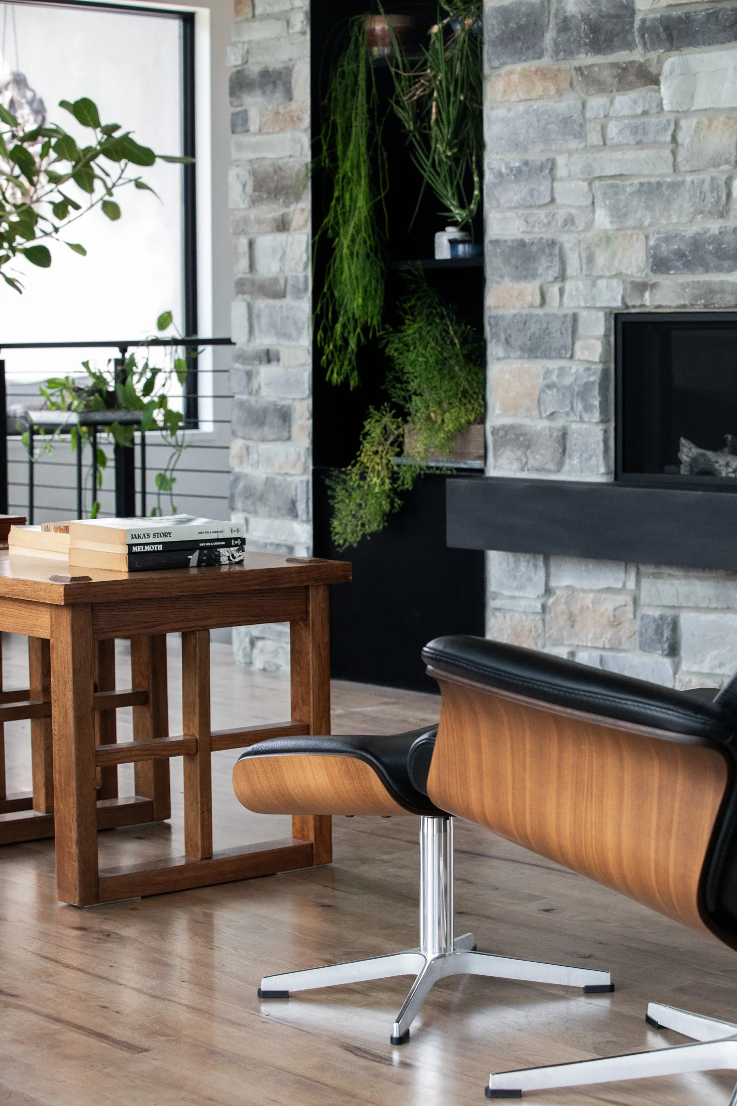 Modern living room with a stone fireplace, wooden coffee table, black and wooden lounge chair, indoor plants, and a large window. Interior Design photography.